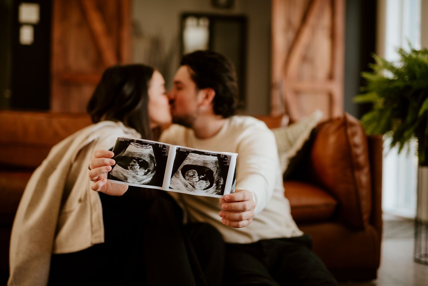 Couple sharing intimate moment while holding ultrasound photos on couch.