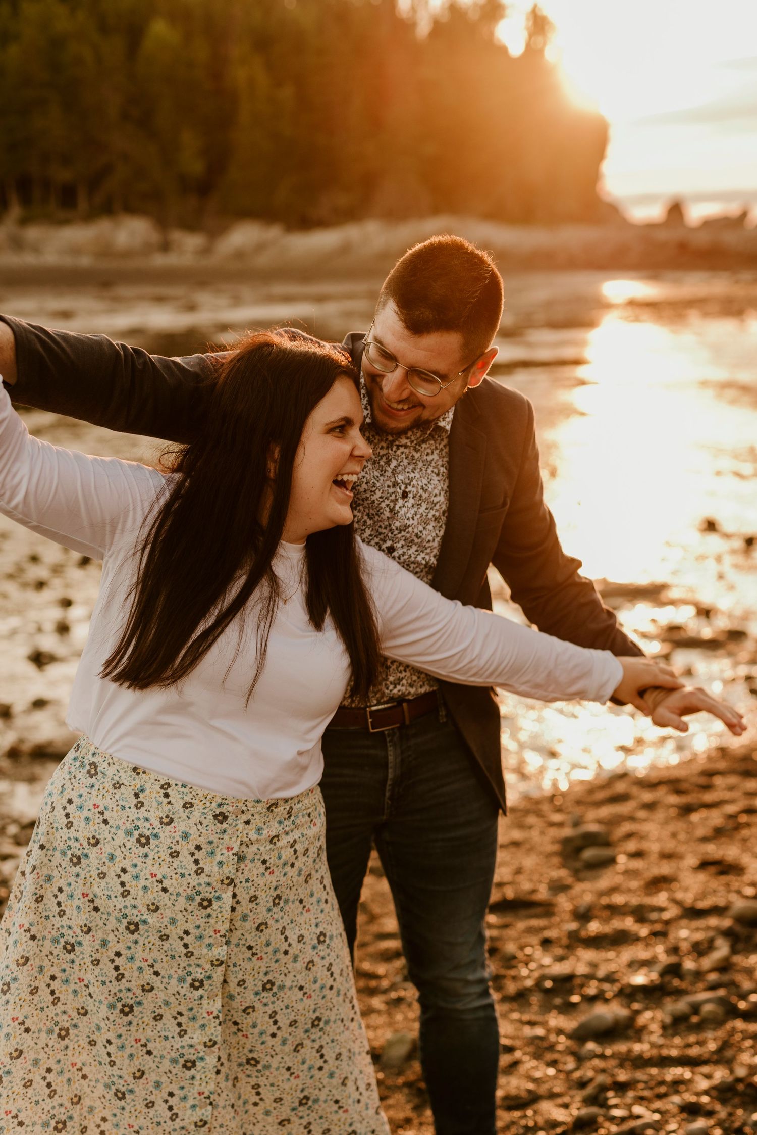 Couple laughing and dancing together on rocky beach at sunset.