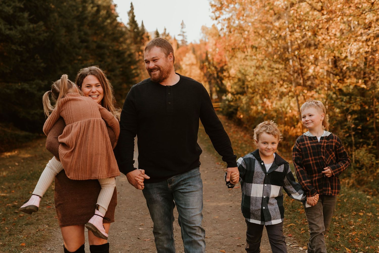 Family of four walking on autumn trail, parents holding children's hands, surrounded by colorful fall foliage.