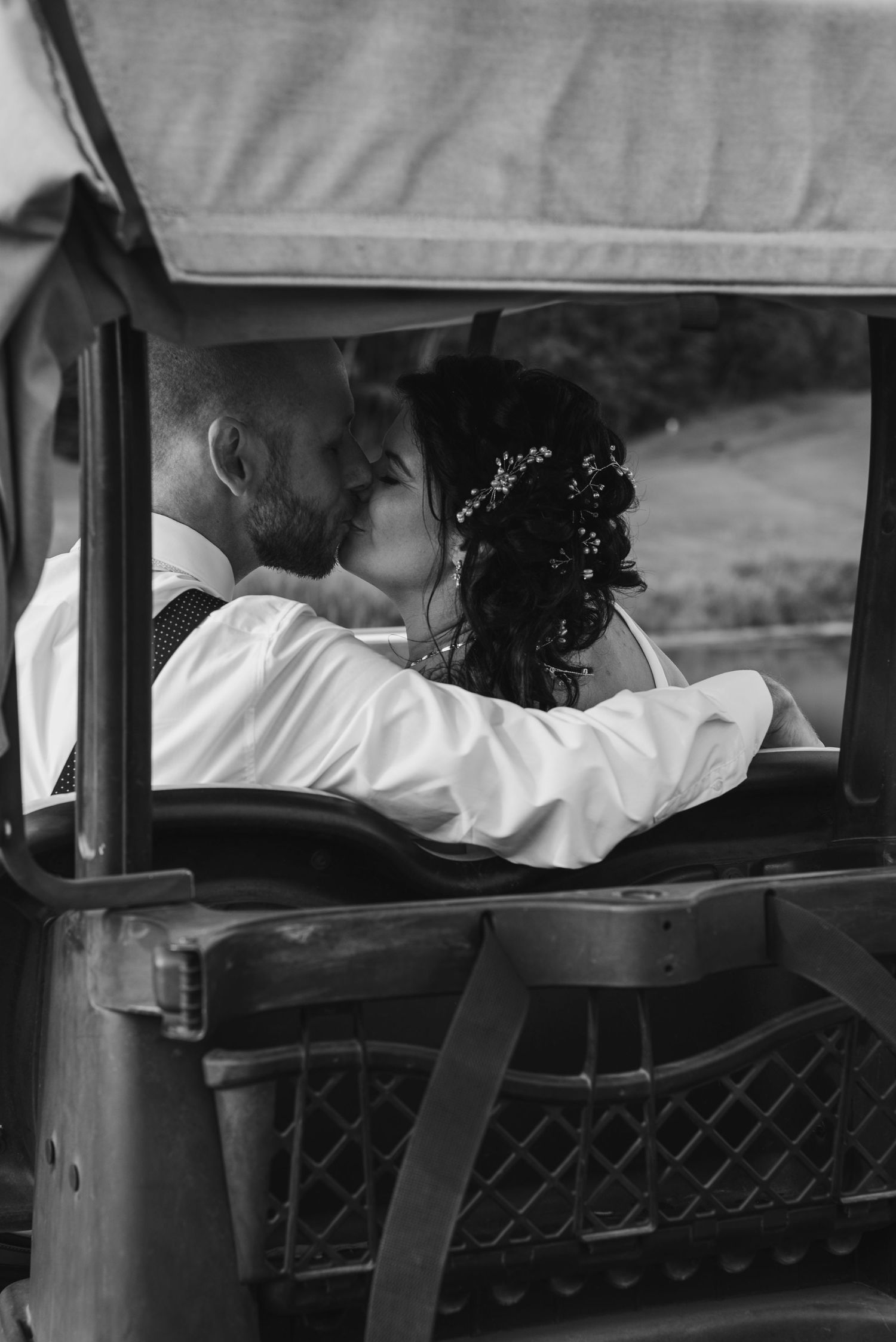 Un couple de jeunes mariés, installé à l'arrière d'une voiture d'époque, s'embrasse tendrement, le tout capté en noir et blanc.