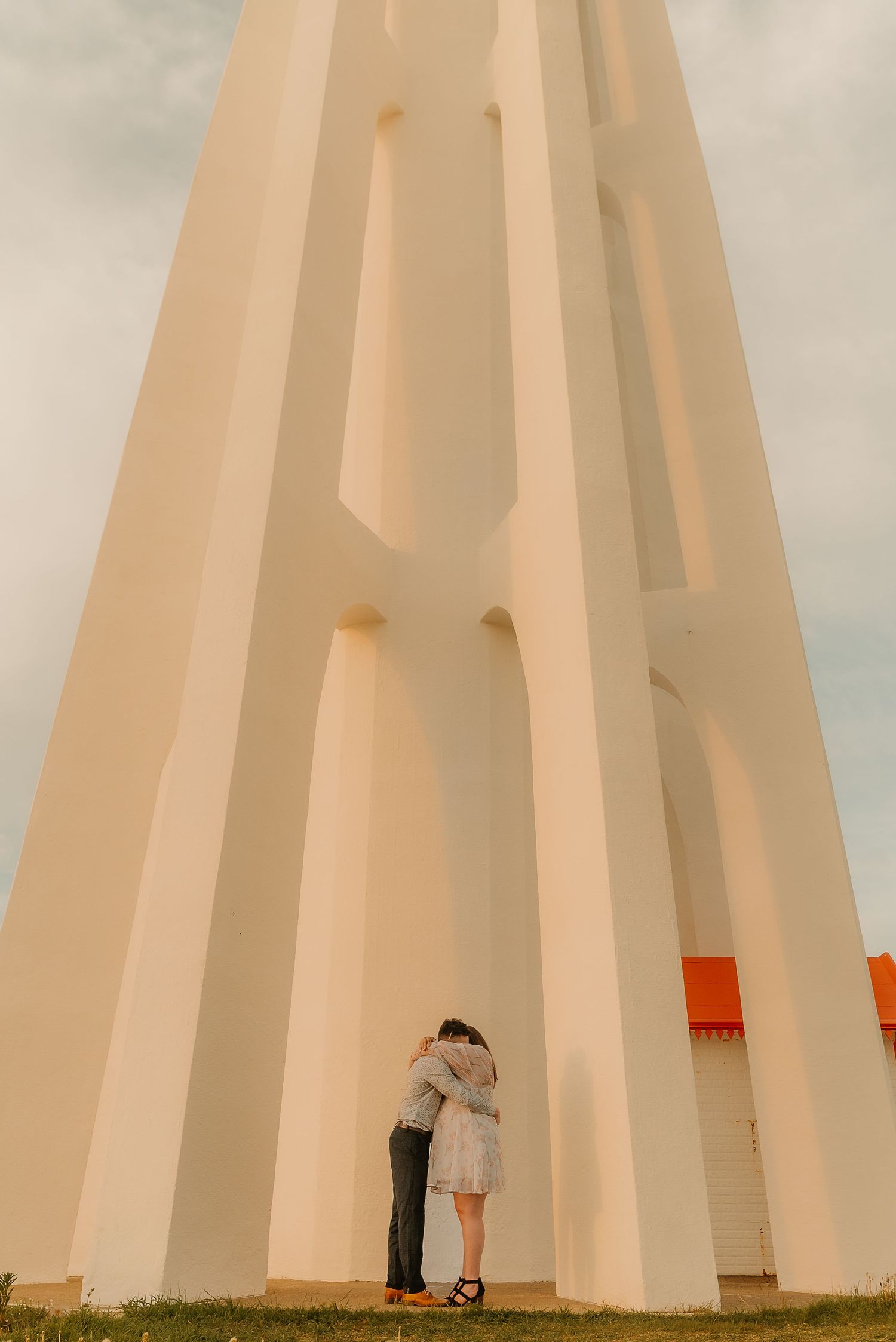 Couple embracing at the base of a large white architectural monument under a cloudy sky.