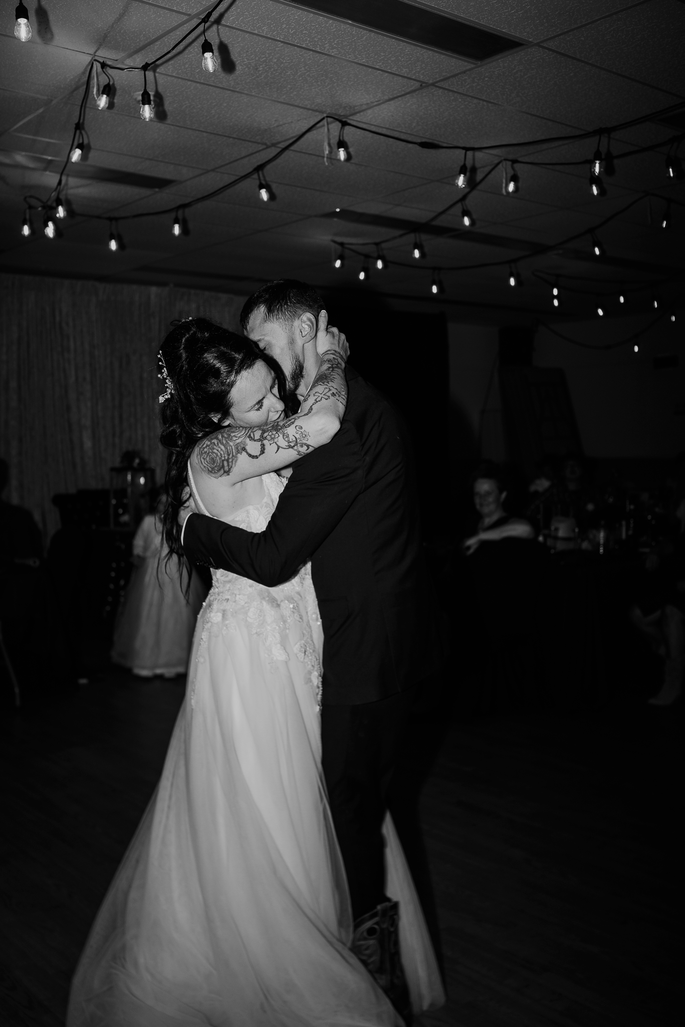 Black and white photo of bride and groom sharing their first dance under string lights at wedding reception.