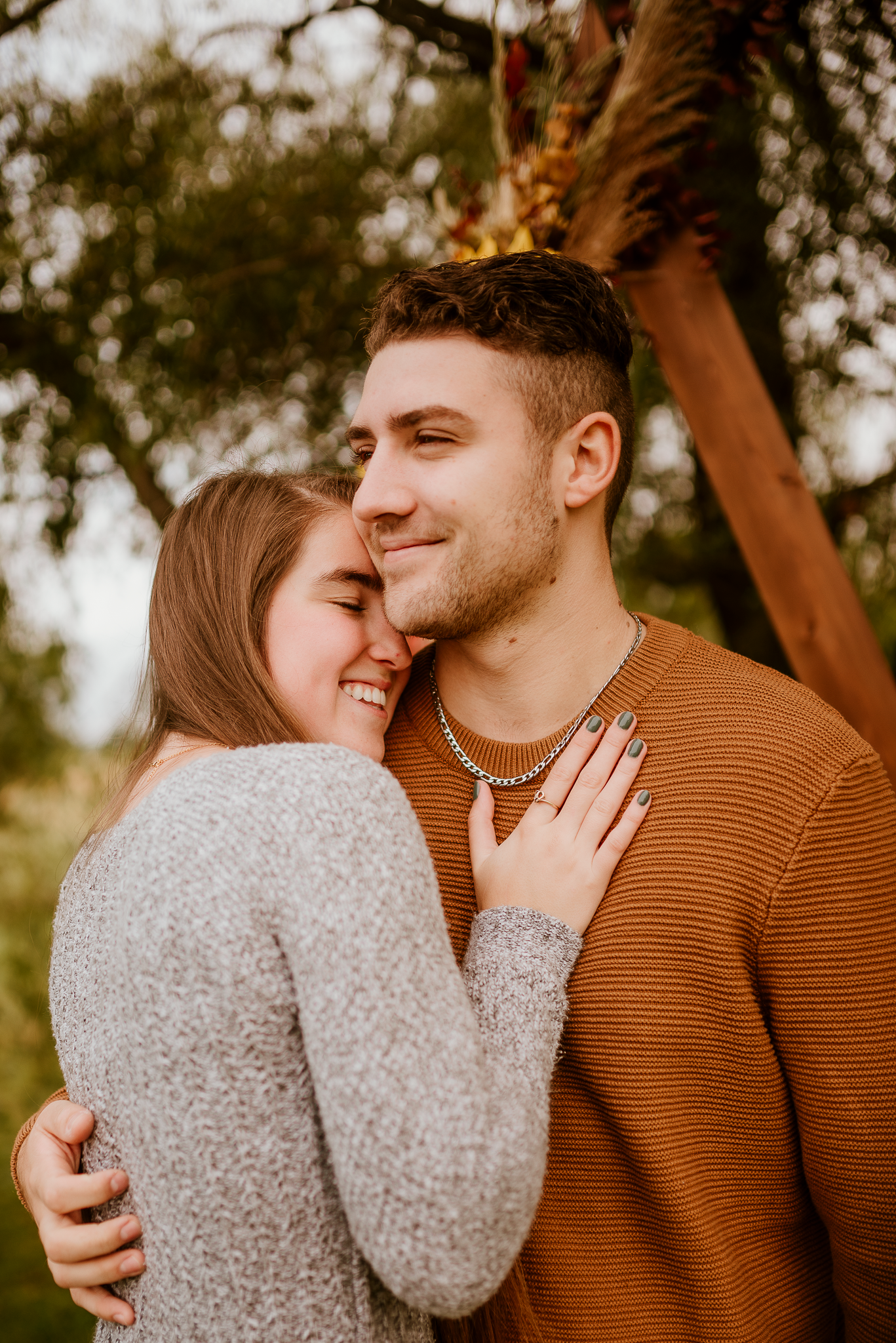 A couple embraces outdoors, woman smiling against man's cheek, man wearing orange sweater and chain necklace.