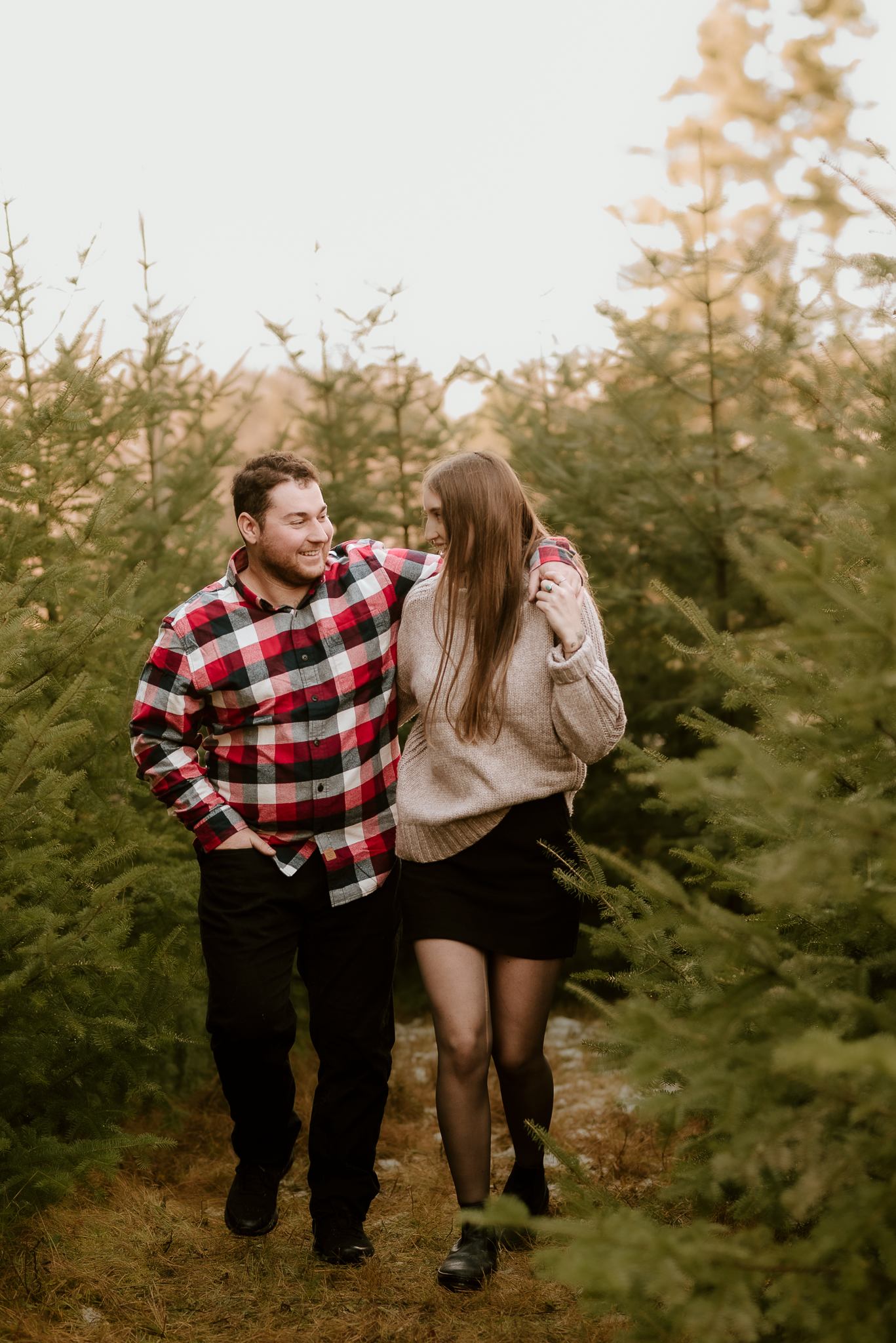 Couple walking through Christmas tree farm, man in plaid shirt and woman in beige sweater smiling at each other.