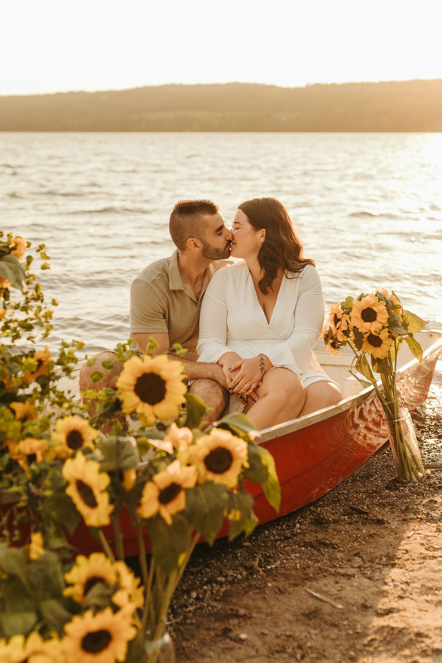 Couple kissing in a red boat surrounded by sunflowers on a lakeside beach at golden hour sunset.