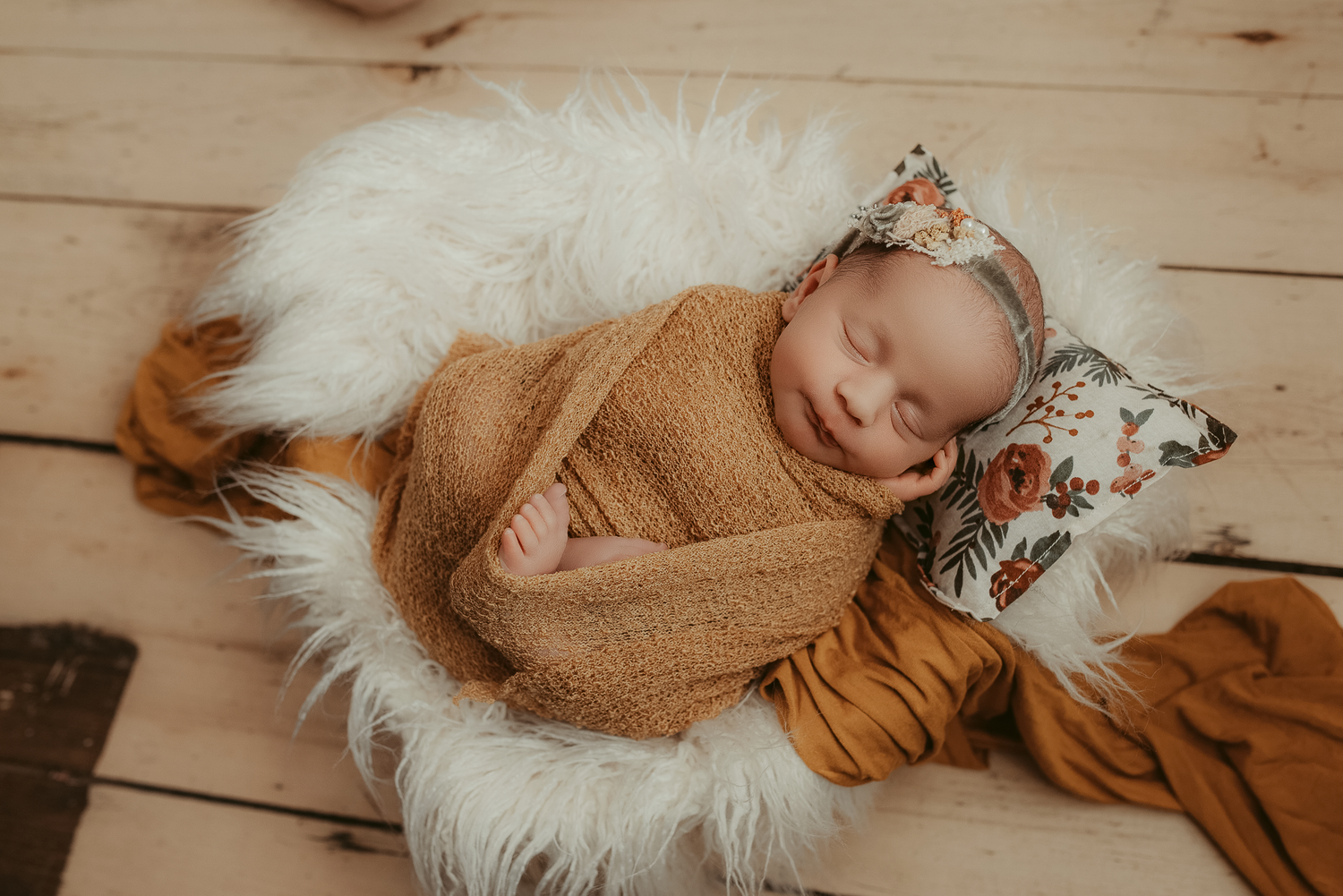 Newborn baby sleeping peacefully in a fur bowl, wrapped in a mustard knit blanket with a floral headband.