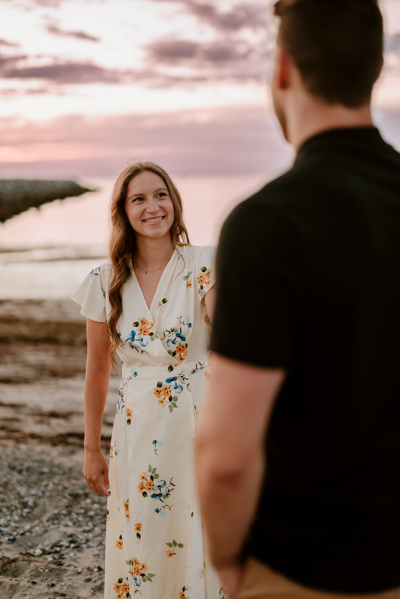 A woman in a floral maxi dress smiles at a man during a romantic beach sunset engagement photo session.