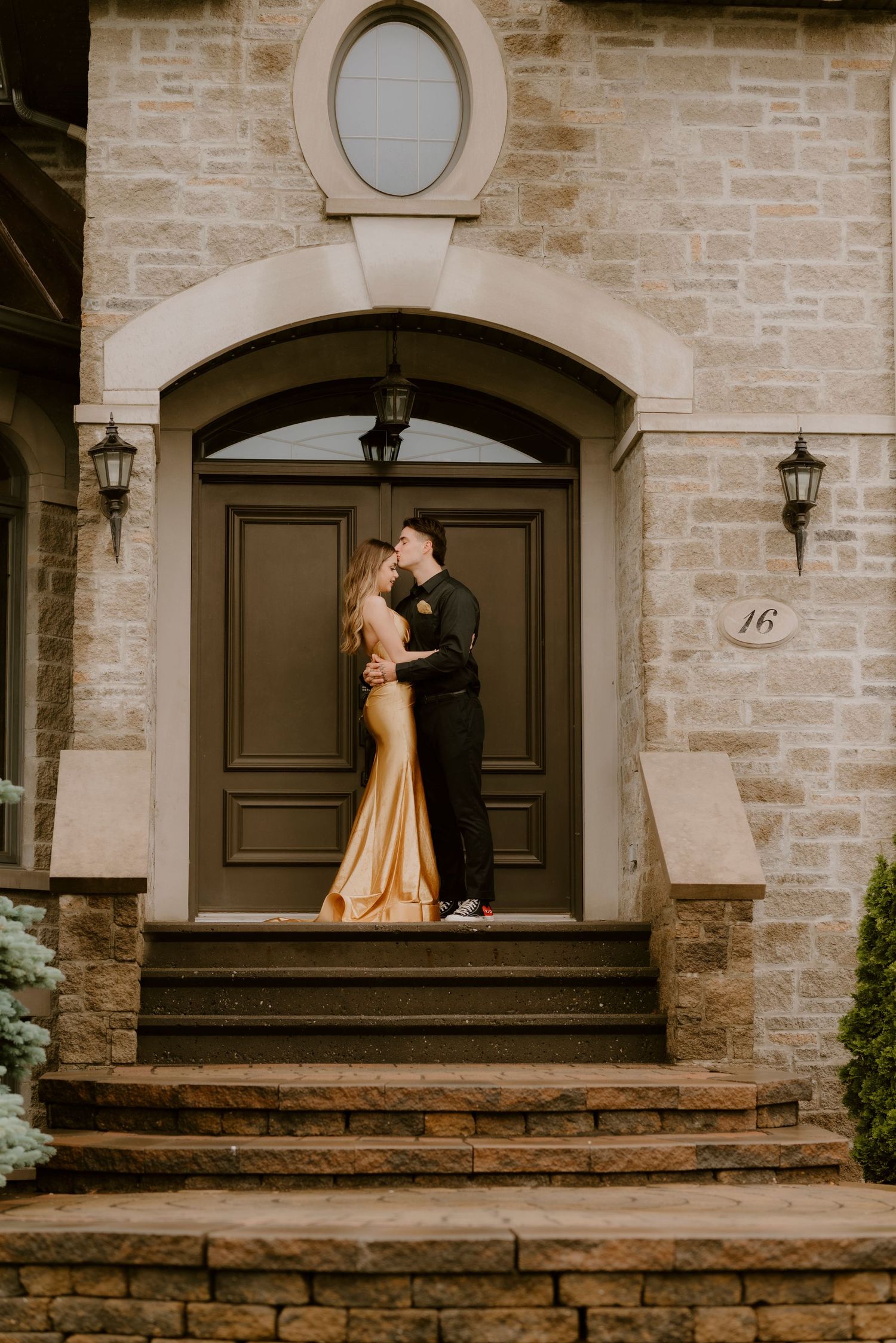 Couple in formal wear kissing on stone steps of elegant home, woman in gold gown, man in dark suit.