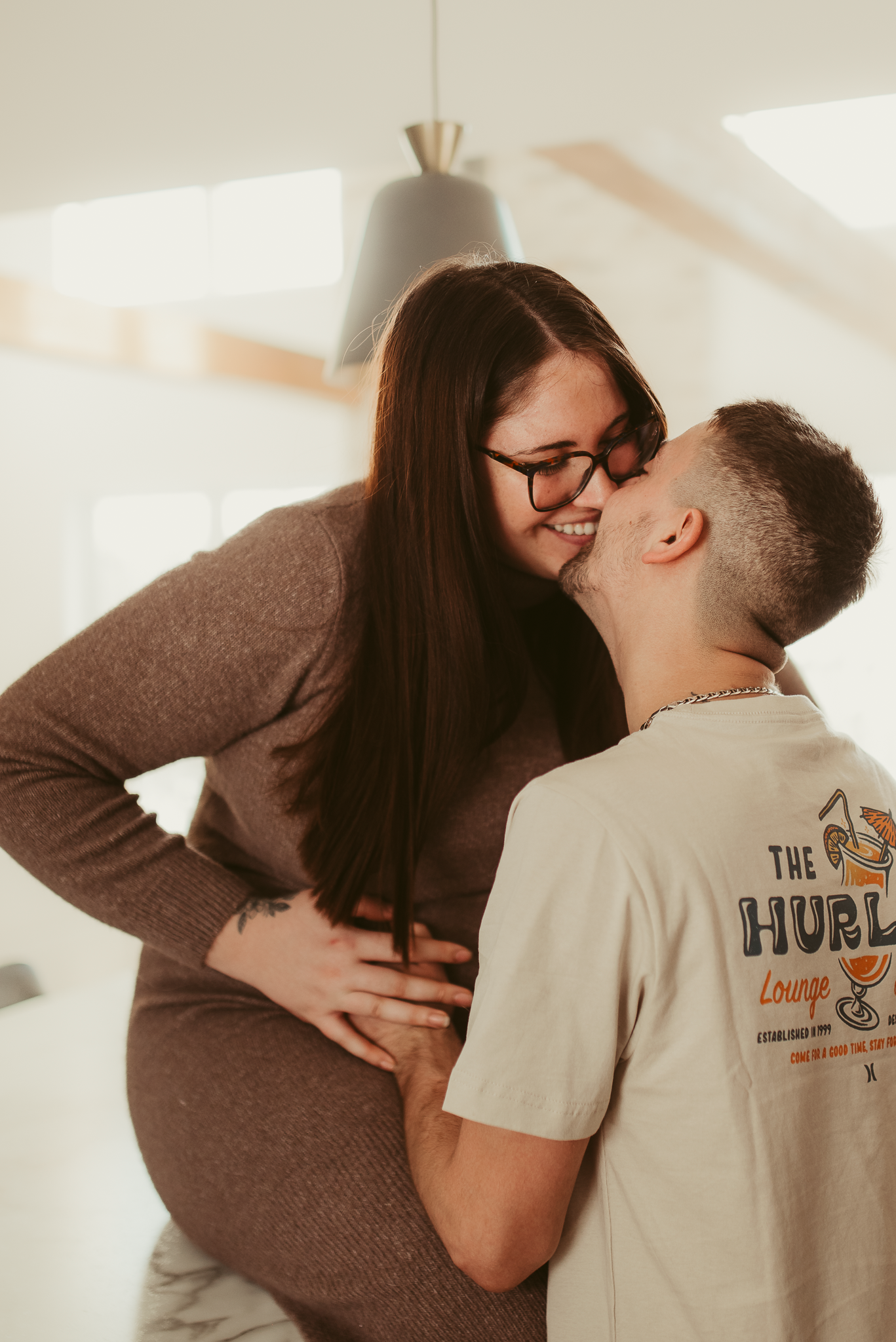 A pregnant woman in a brown dress shares a tender kiss with her partner wearing a white graphic tee indoors.