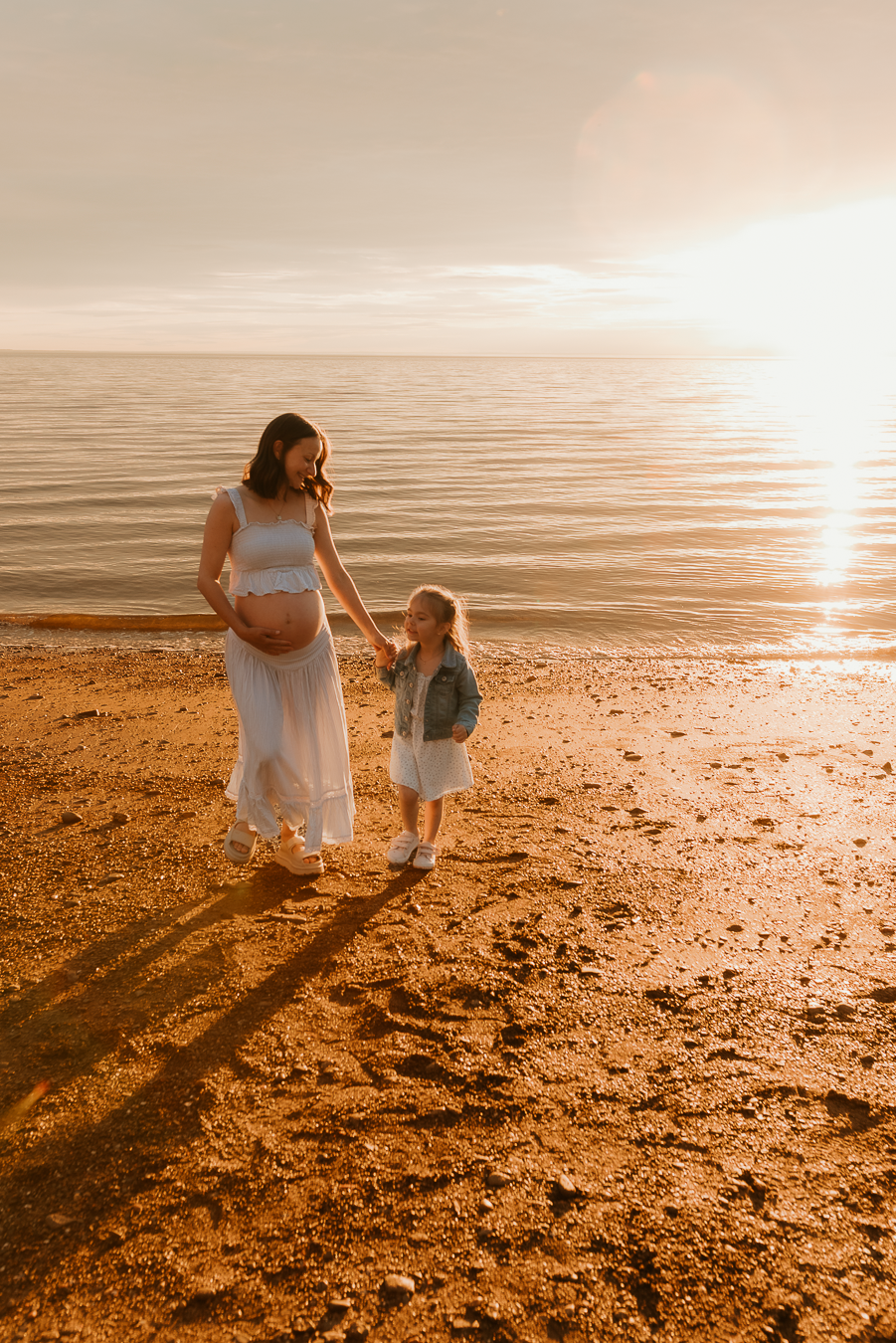 Pregnant woman in white dress holds hands with young child on sandy beach at golden sunset.
