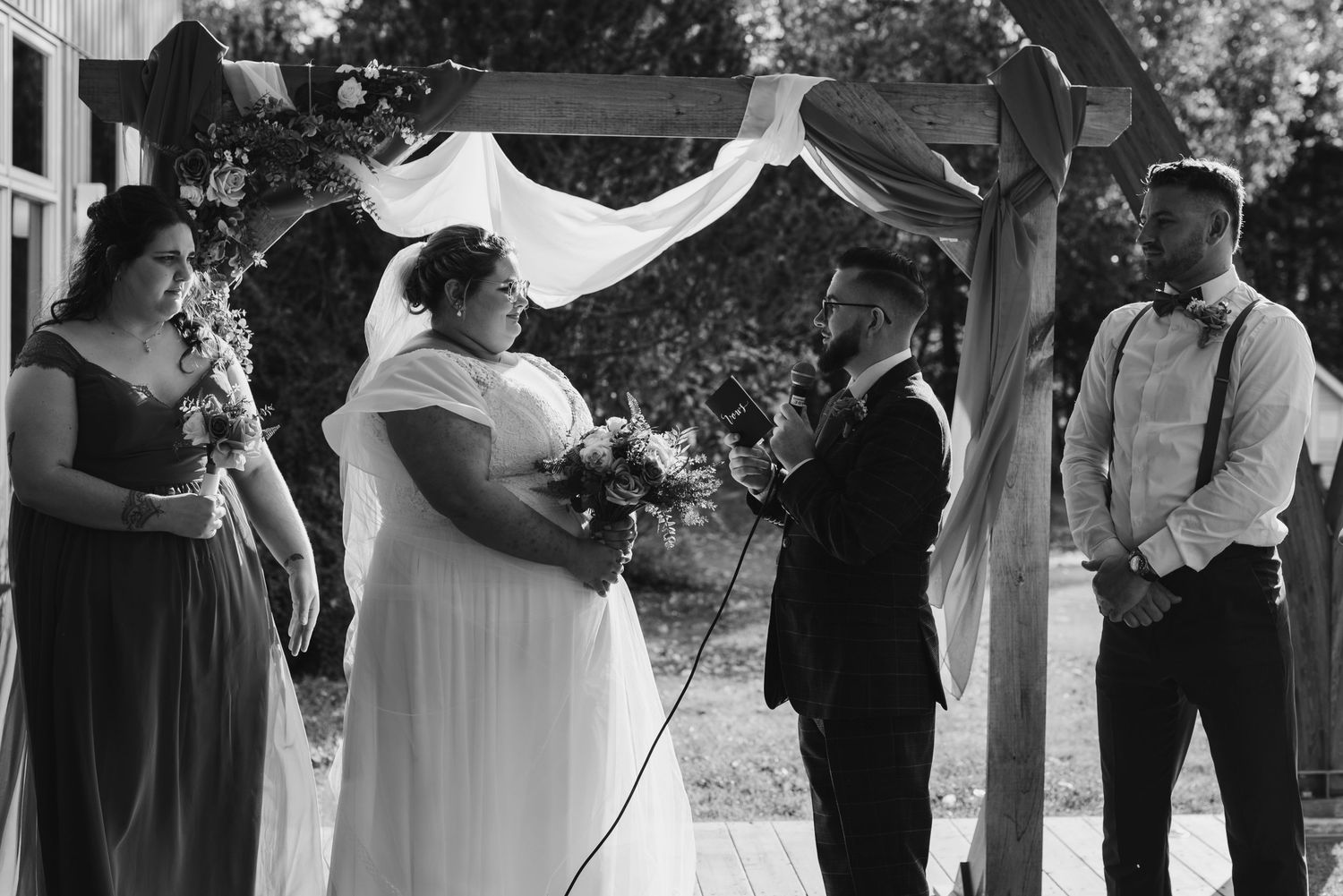 Outdoor wedding ceremony under floral arch with wedding party in formal attire.