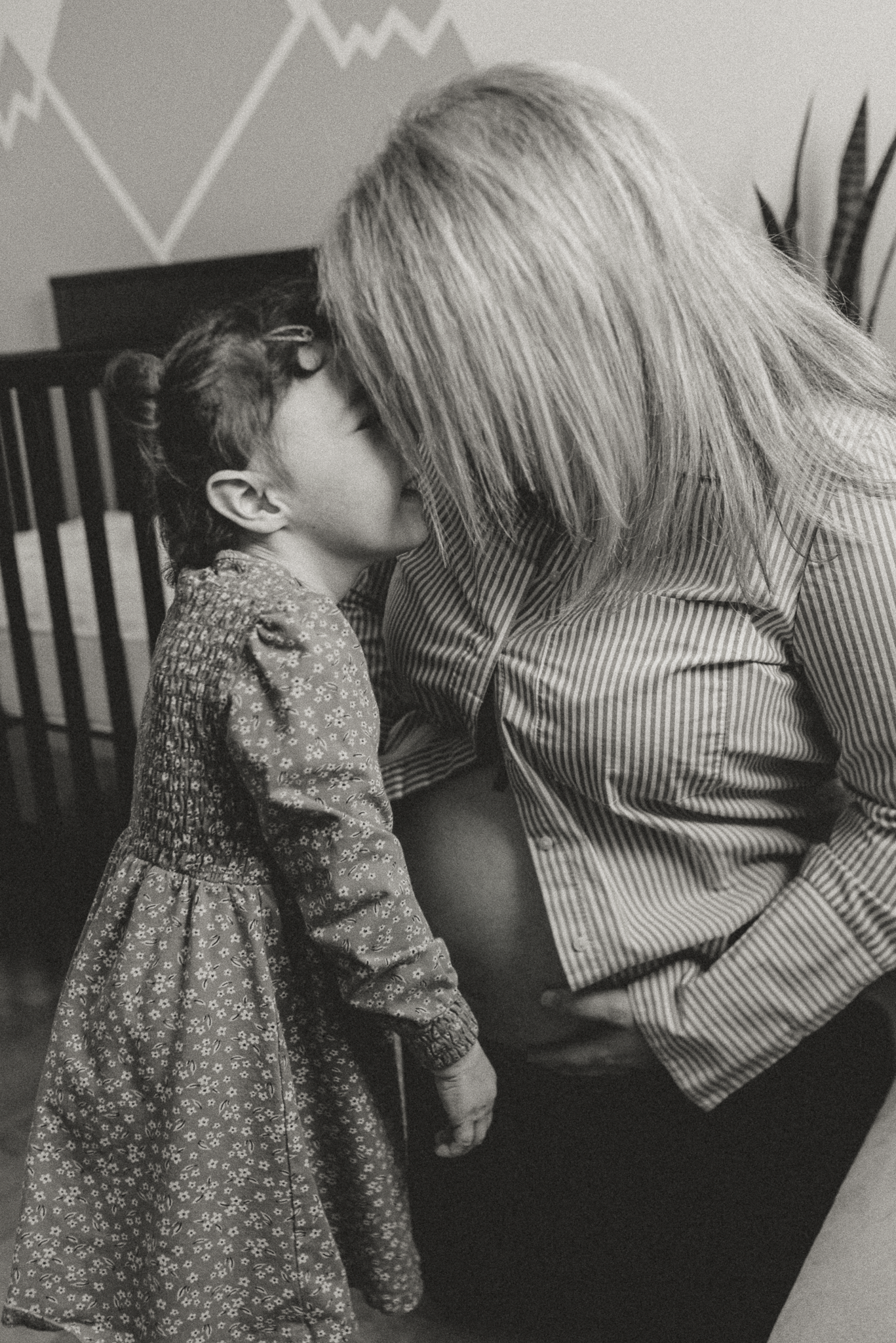 A young girl in a floral dress kisses a pregnant woman's belly in a black and white nursery photo.