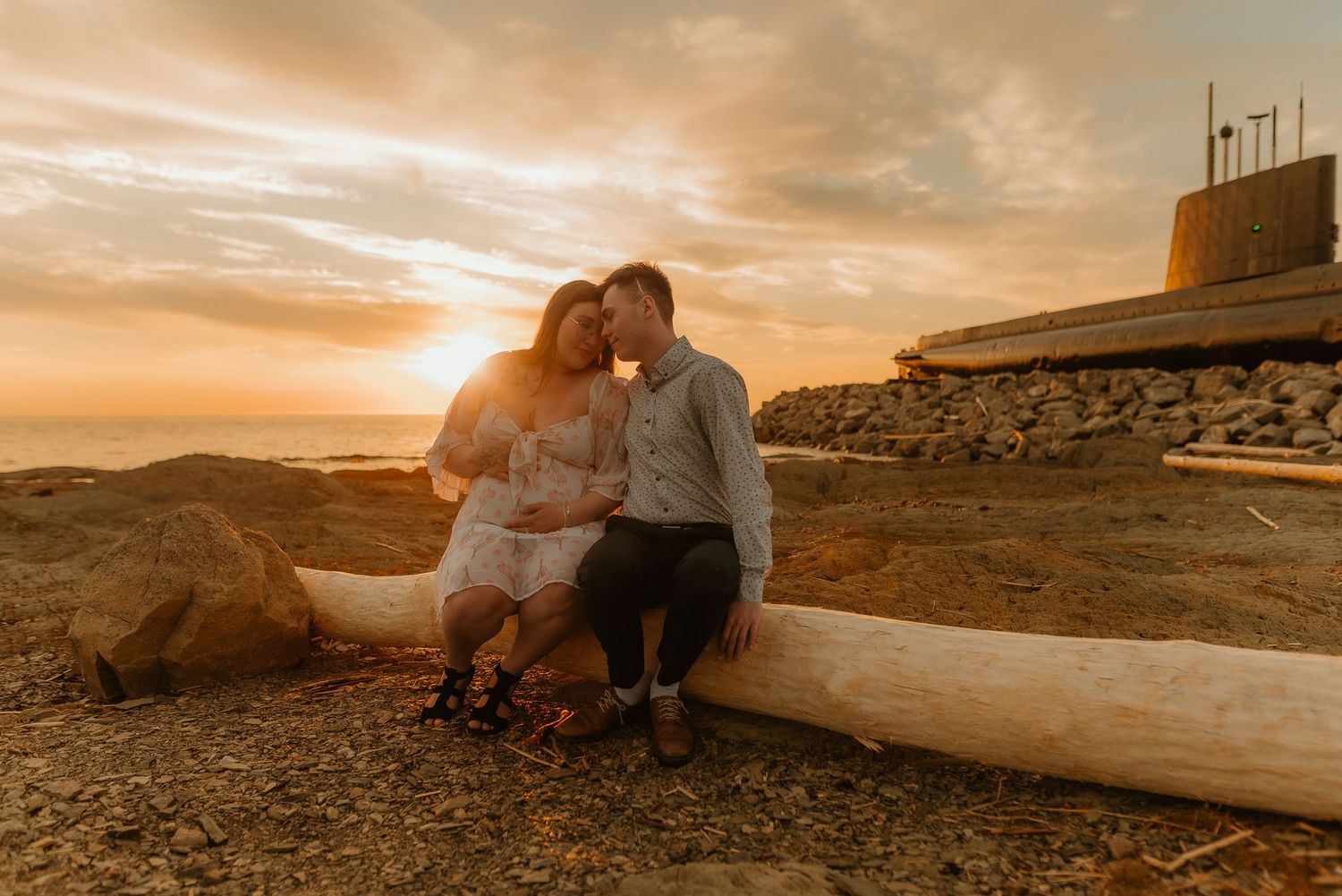Couple sitting on driftwood at golden hour sunset on a rocky beach, sharing a tender romantic moment.