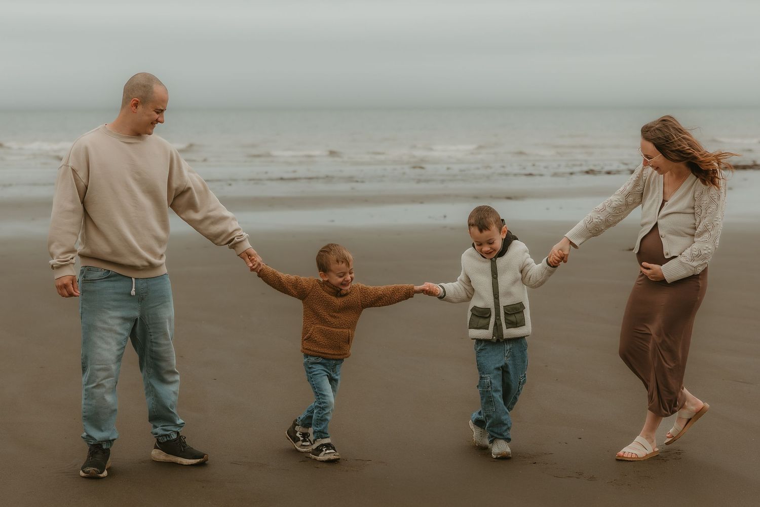 Family of four holding hands on a cloudy beach, two parents swinging their young children between them.