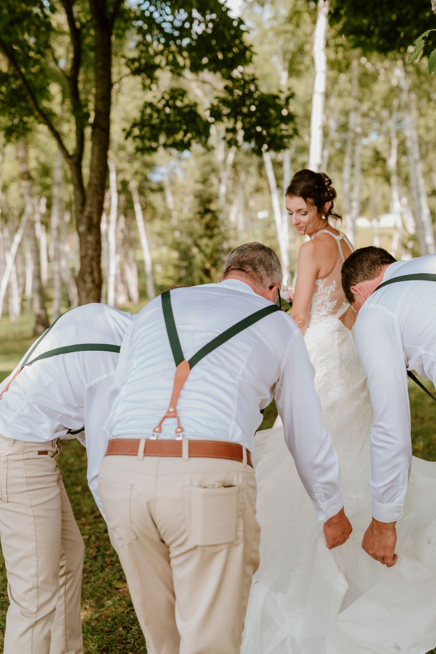 Groomsmen in white shirts and suspenders bow down in a playful outdoor wedding photo pose.