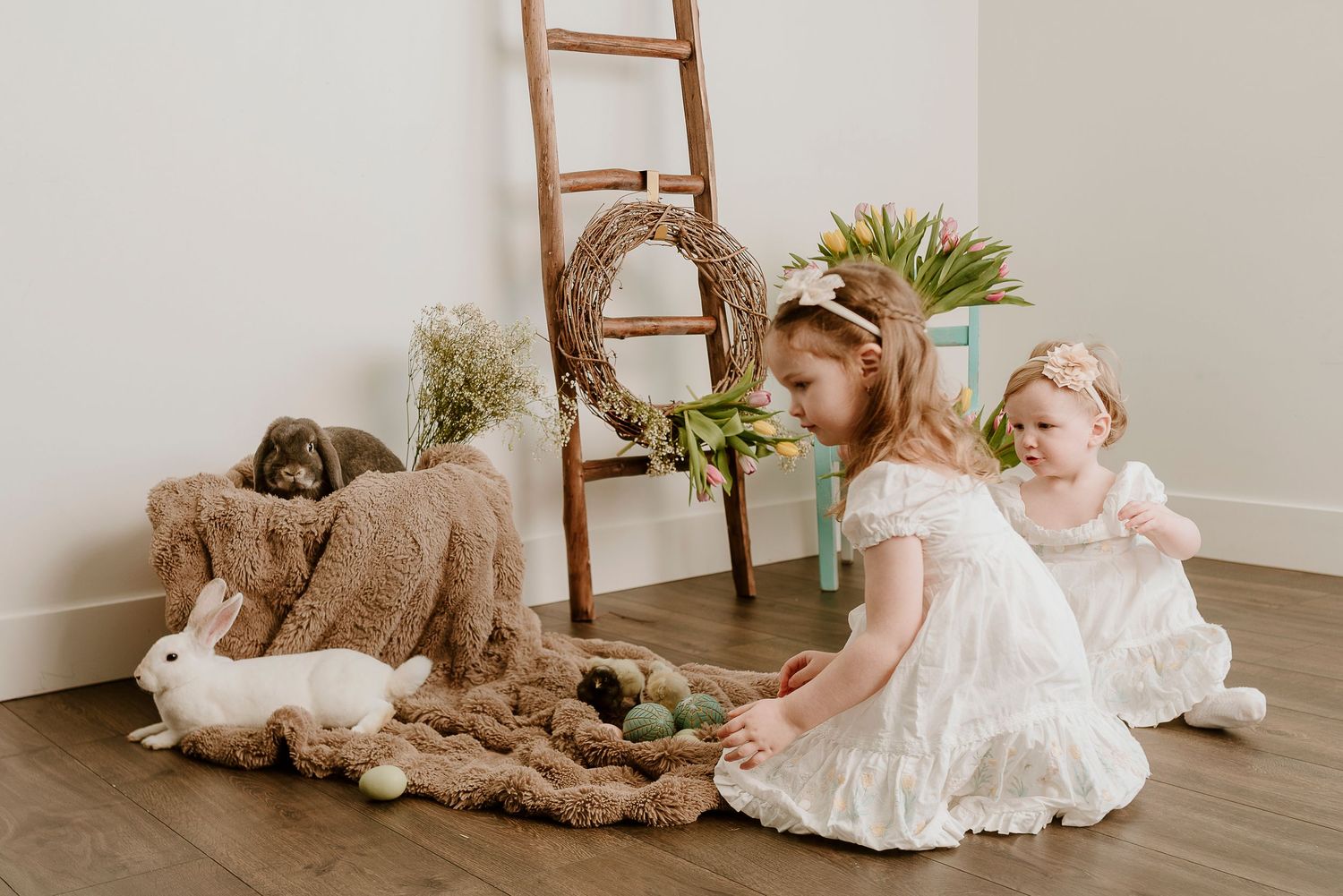 Two young girls in white dresses play with Easter eggs and bunny decorations on a fuzzy blanket indoors.