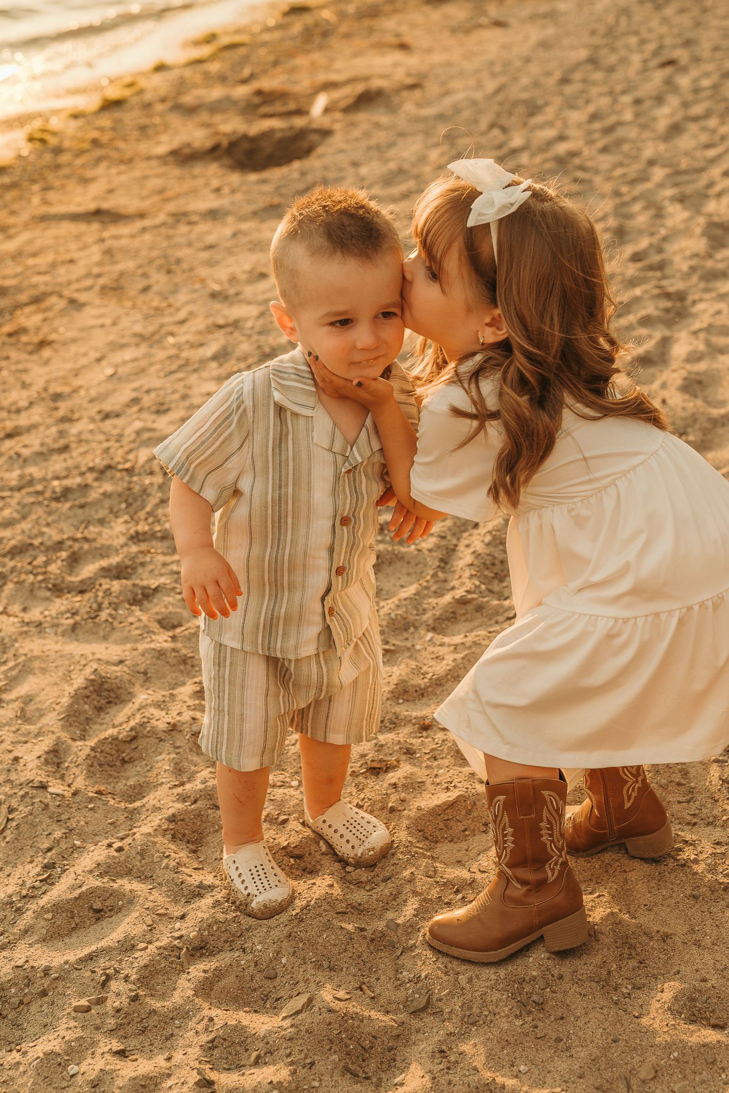 A young girl in a white dress kisses a toddler boy on the cheek on a sandy beach at golden hour.