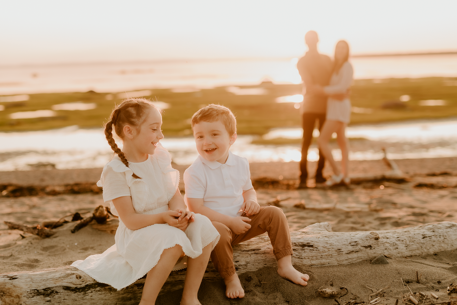 Two young children laugh and sit on beach sand at golden hour, with their parents embracing in the background.