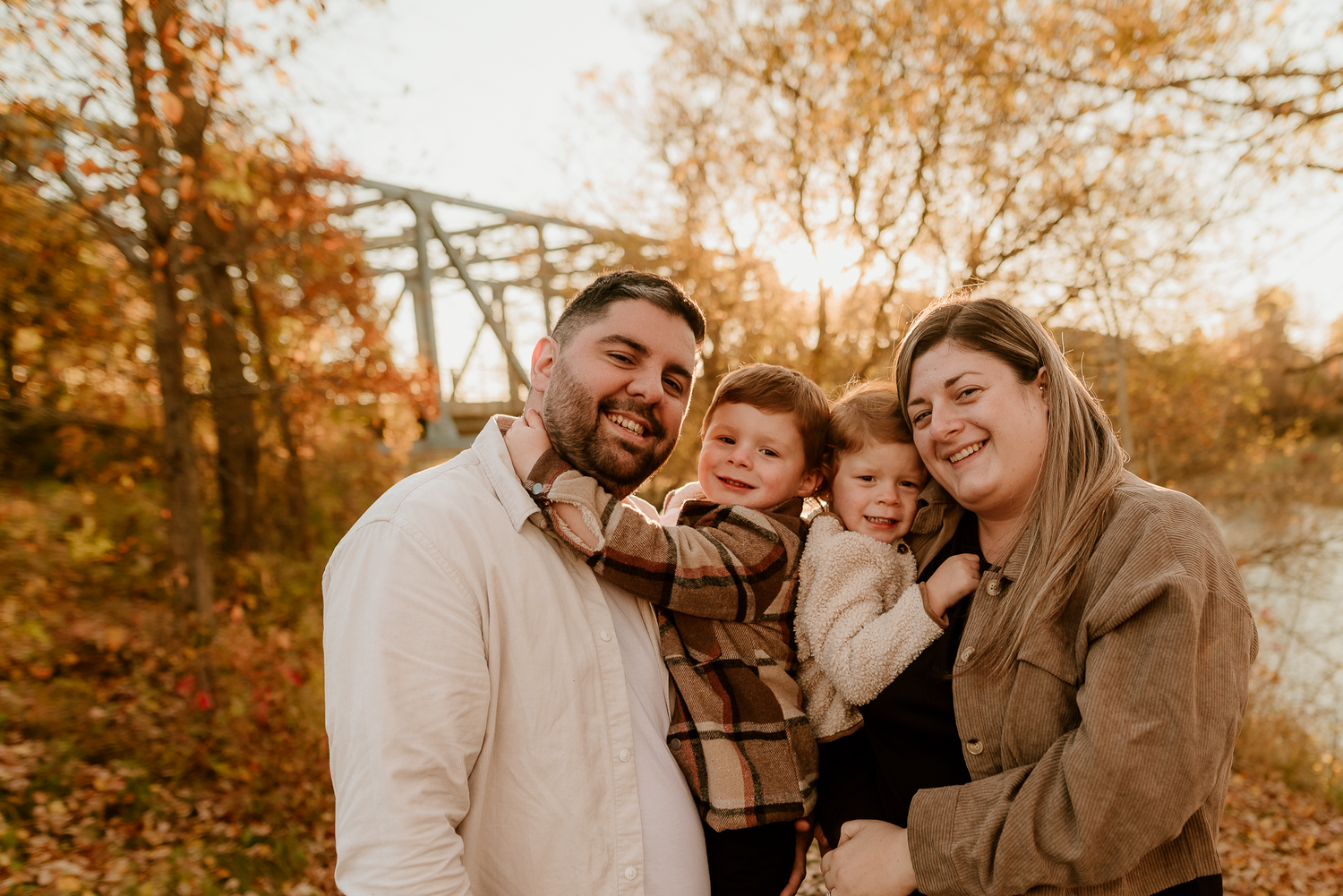 Happy family of four smiling outdoors in autumn, parents holding two young children with fall foliage and bridge backdrop.