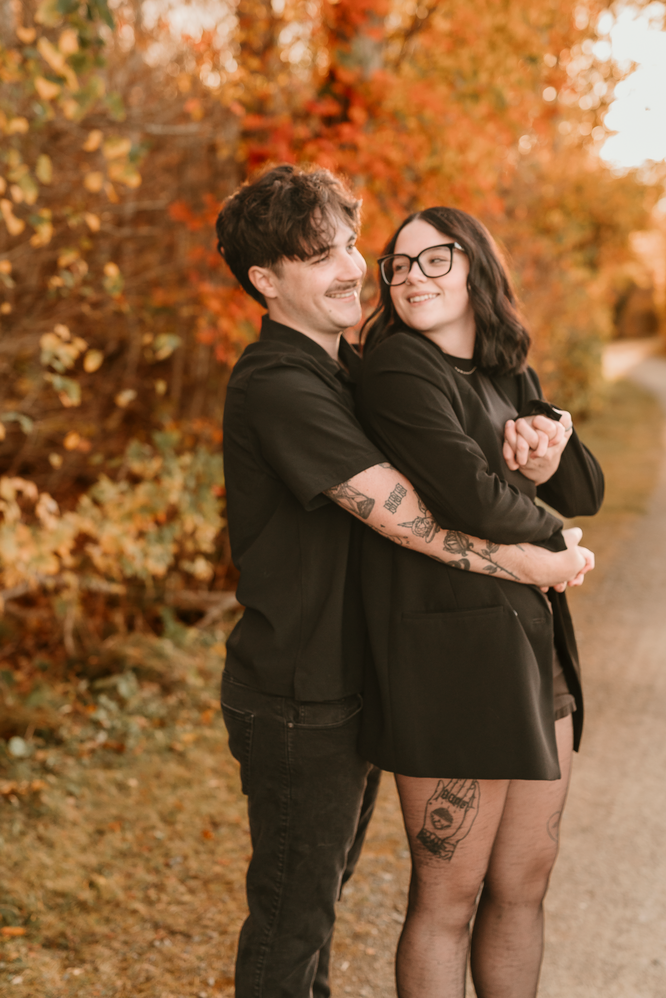 A couple in matching black outfits embraces on a path surrounded by vibrant autumn foliage, both smiling warmly.
