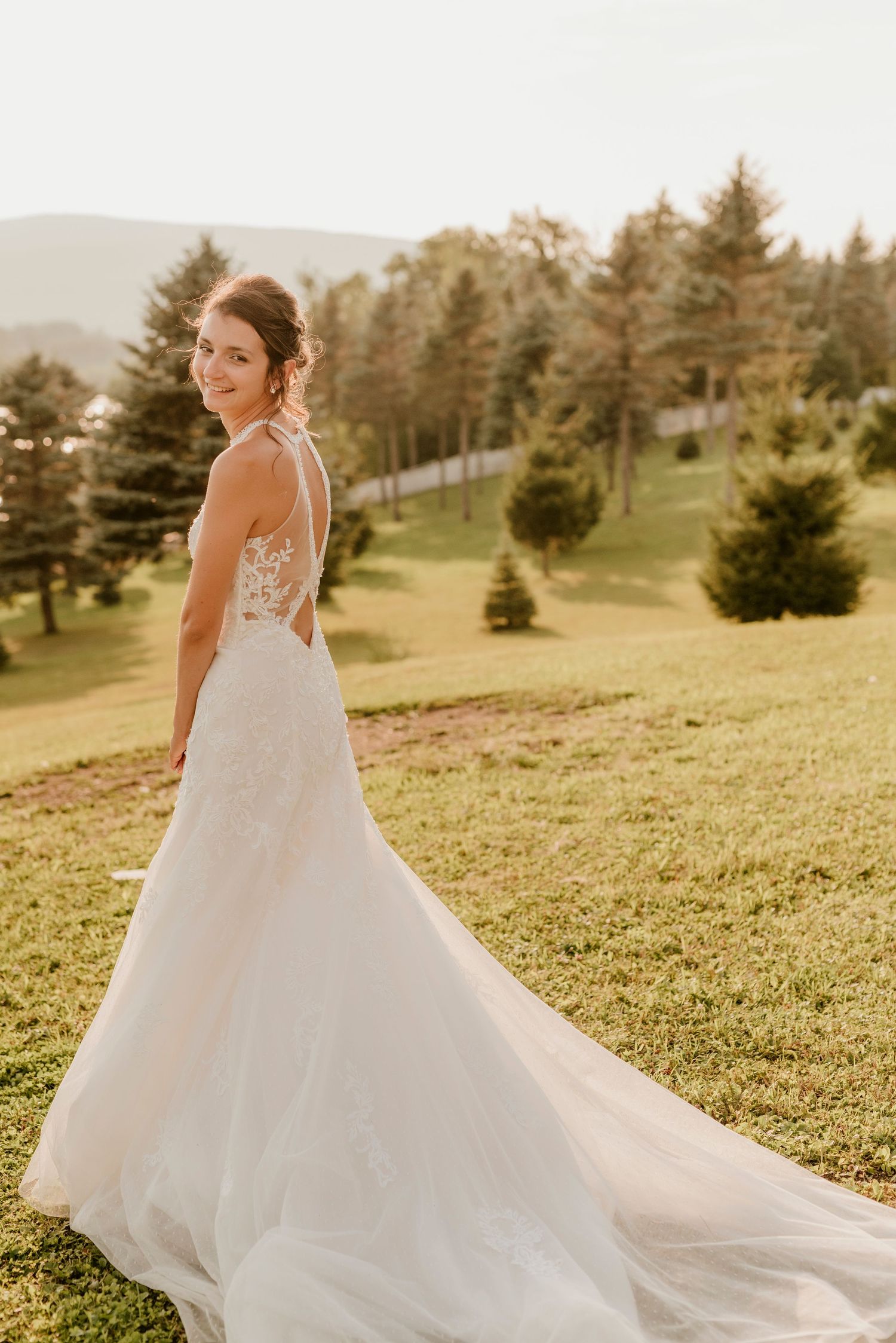A bride in a lace wedding gown poses on a grassy hillside with mountains in the background.