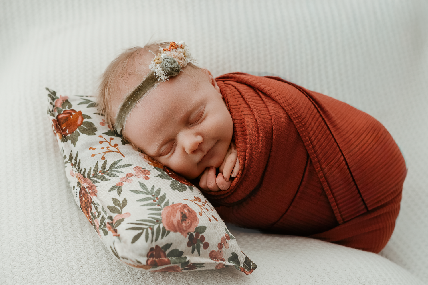 Sleeping newborn baby swaddled in rust orange wrap resting on floral pillow with delicate flower headband.