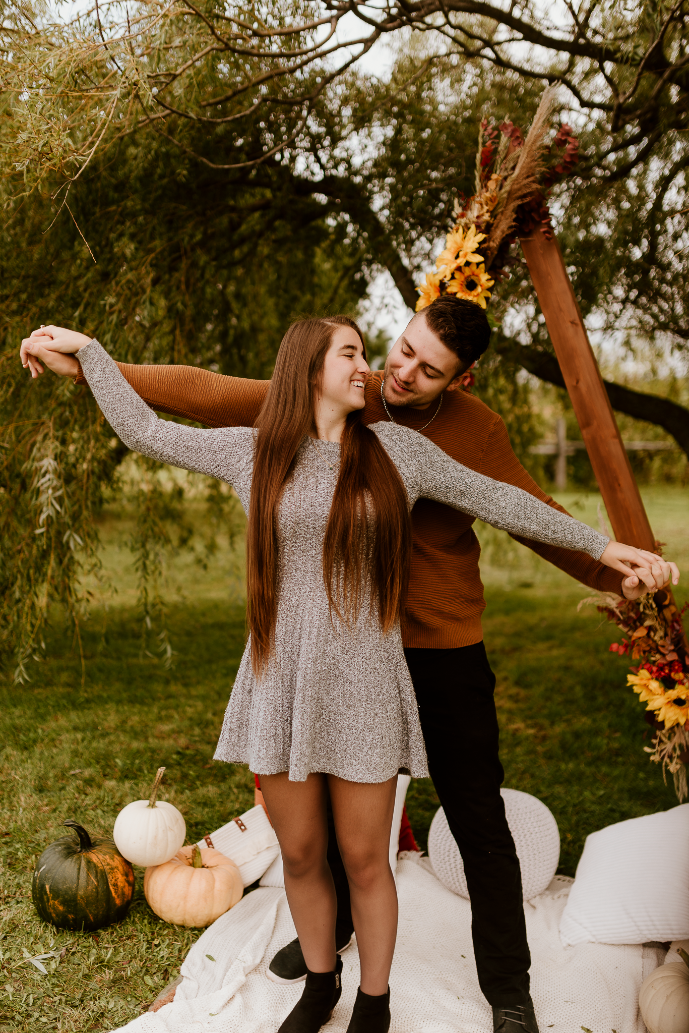 A young couple shares a romantic autumn moment outdoors with pumpkins, fall flowers, and warm earthy tones in a garden setting.