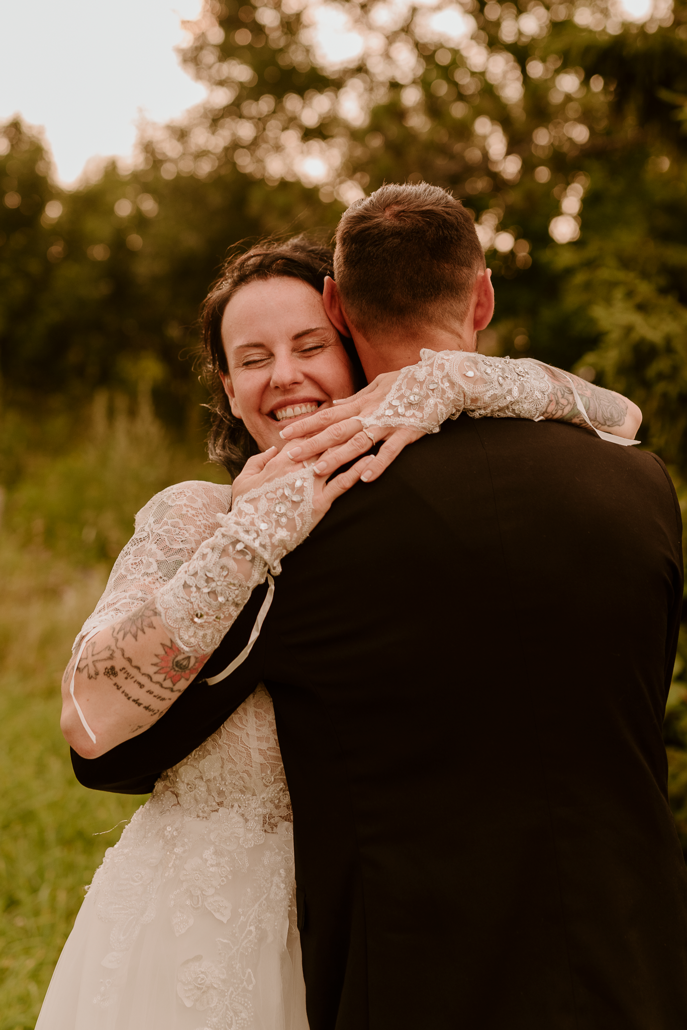 A joyful bride in a lace wedding dress embraces her groom outdoors, smiling warmly during golden hour.
