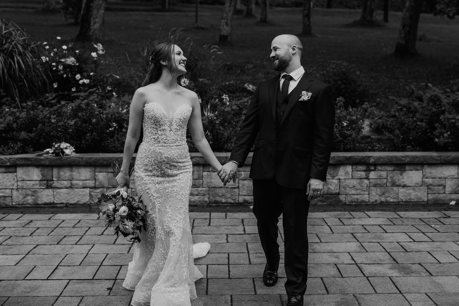 Black and white photo of newlyweds walking hand in hand on stone pathway.