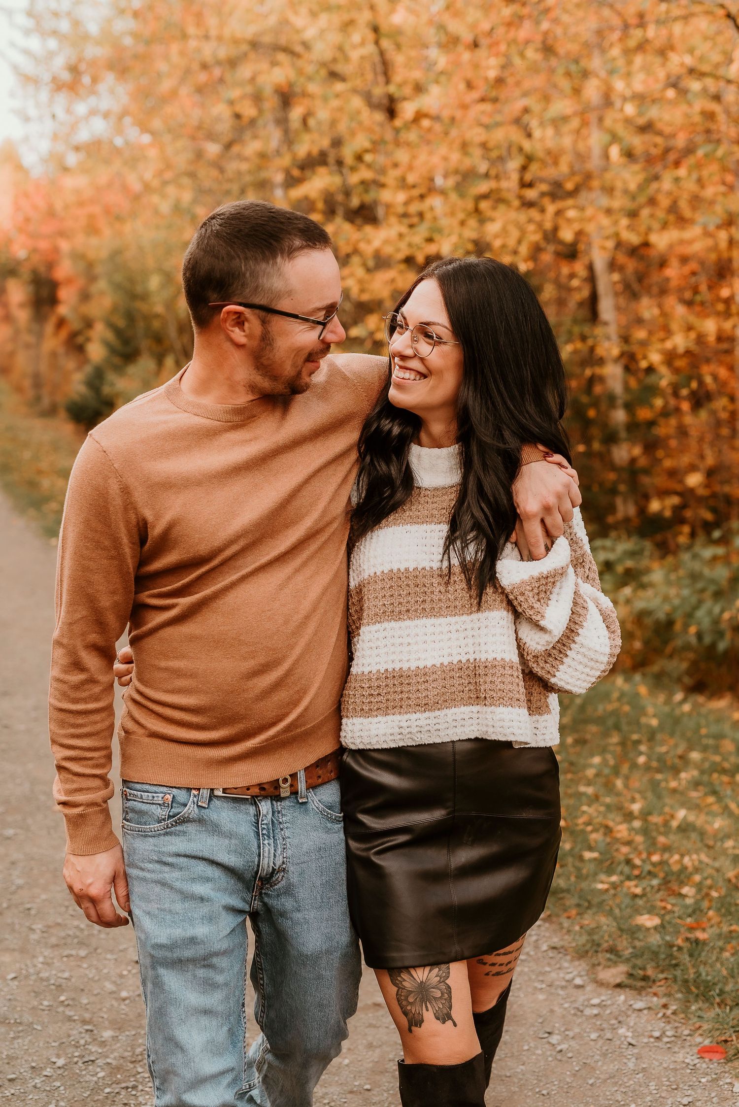 Couple smiling and embracing outdoors in fall foliage, woman in striped sweater and leather skirt, man in tan sweater and jeans.