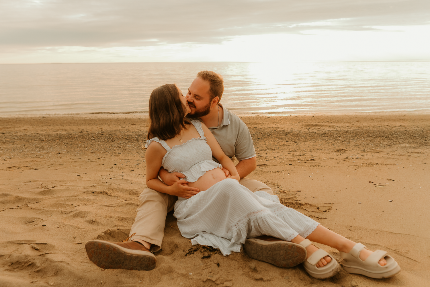 Couple sitting on sandy beach at sunset, man embracing pregnant woman in white dress, romantic maternity photo.