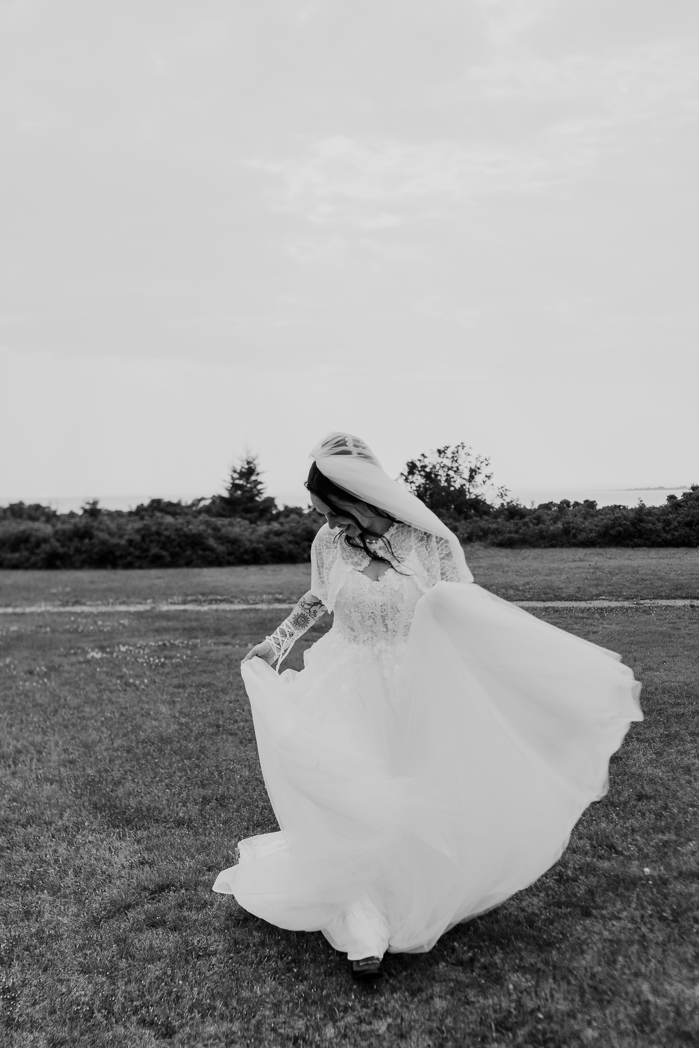 Black and white photo of a bride twirling in a lace wedding gown outdoors on a green lawn.