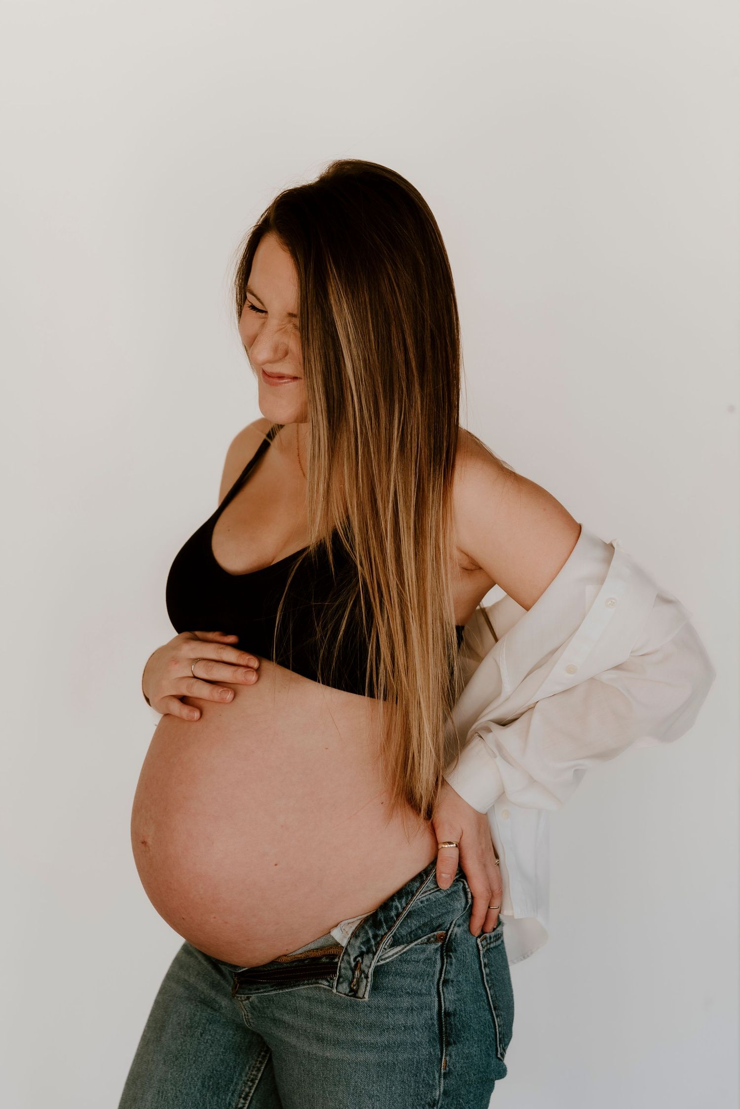 Pregnant woman in black crop top and jeans holding baby bump, wearing open white shirt, posing against white background.
