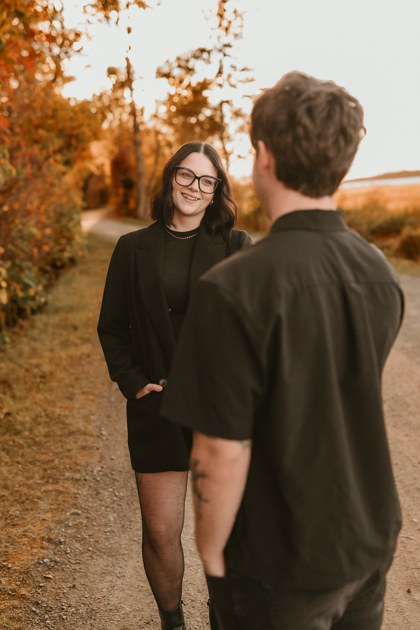 A couple shares a romantic moment on an autumn path, woman in black blazer and glasses smiling warmly at her partner.