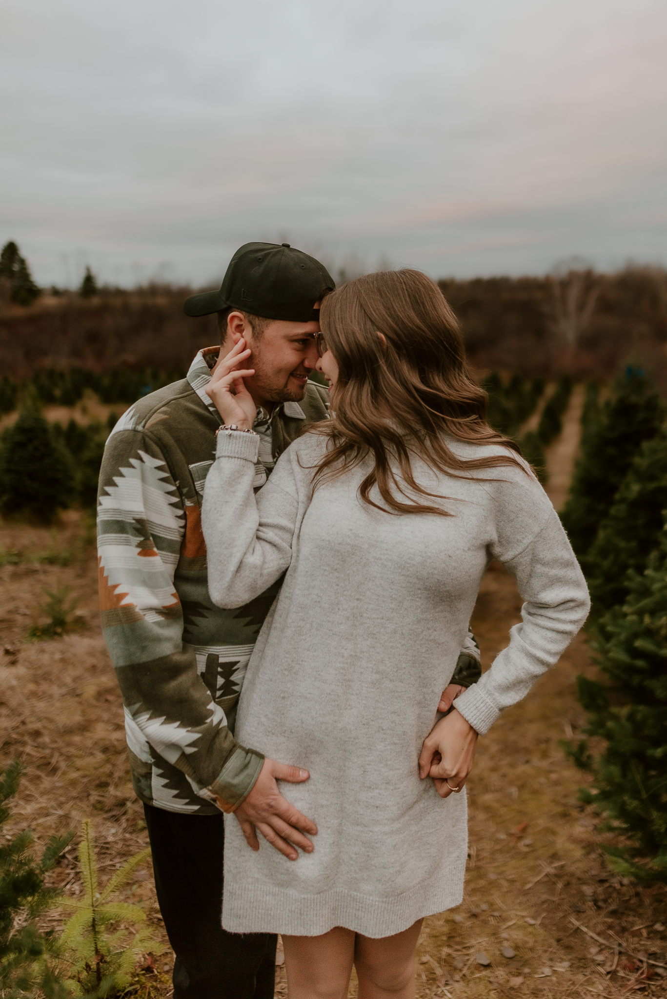 Couple sharing a romantic moment at a Christmas tree farm, woman in gray sweater dress, man in patterned jacket and cap.