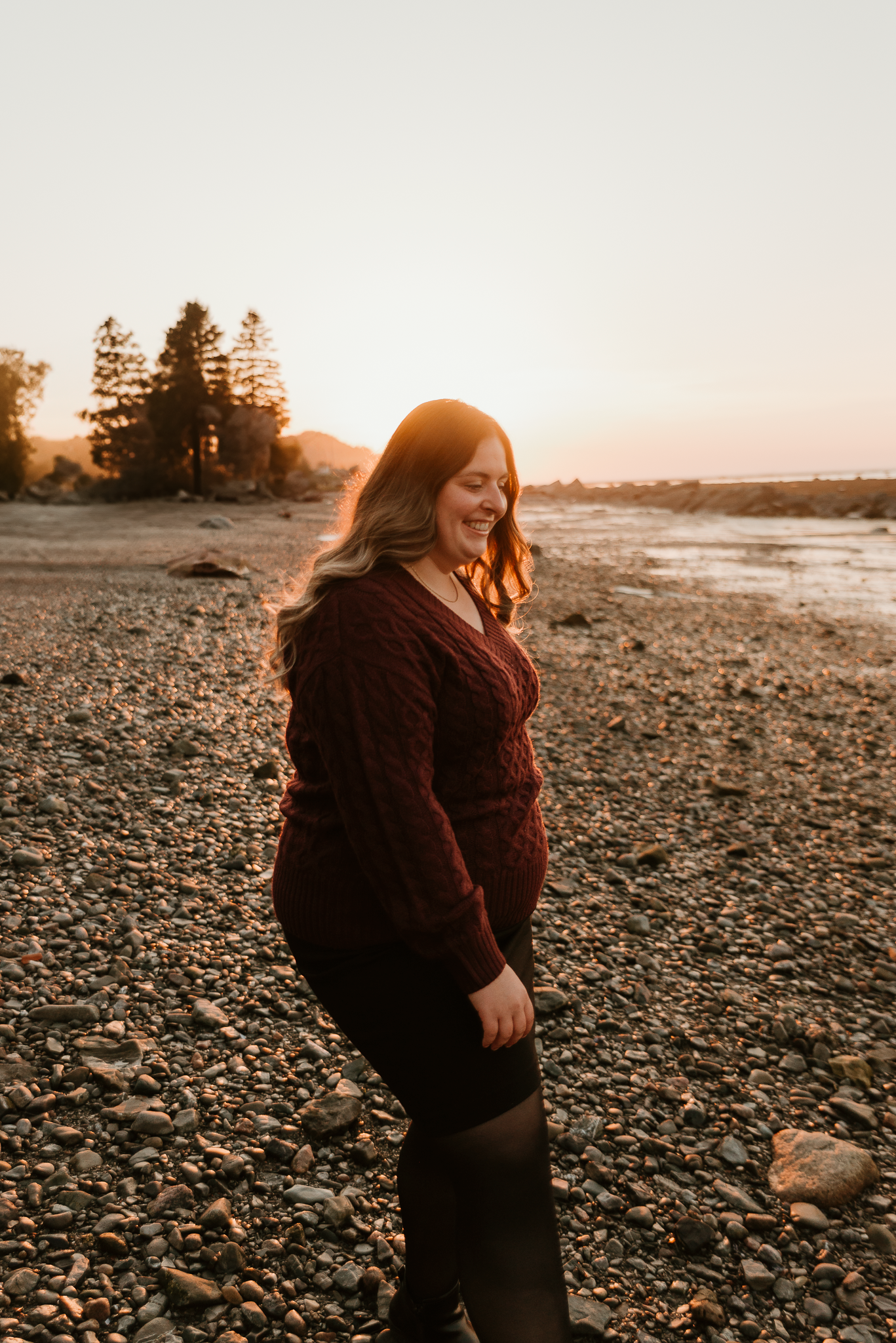 Woman in burgundy cable-knit sweater smiling on rocky beach at golden sunset with trees in background.