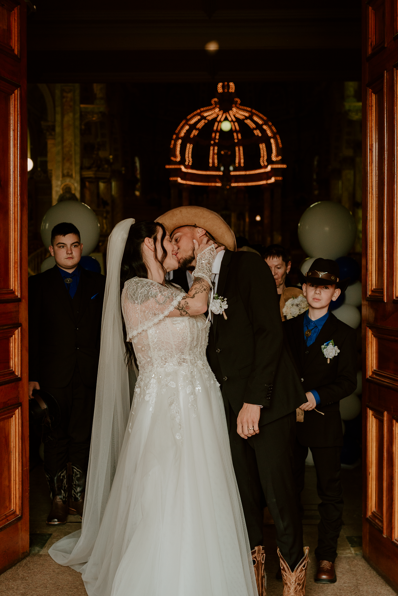 Bride and groom share first kiss at rustic wedding venue, groom wearing cowboy hat, children in blue suits nearby.