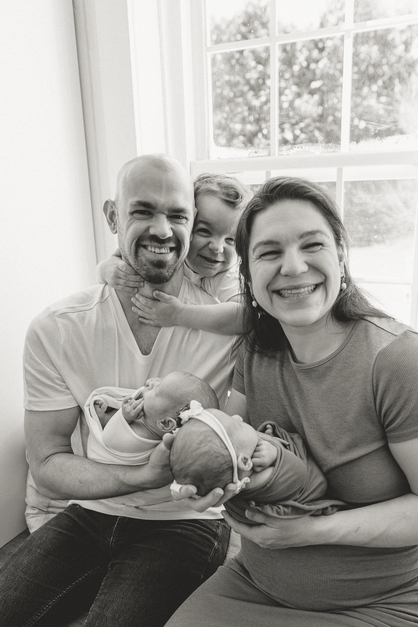 Black and white family portrait with parents smiling, holding newborn baby, and young child peeking from behind, near window.