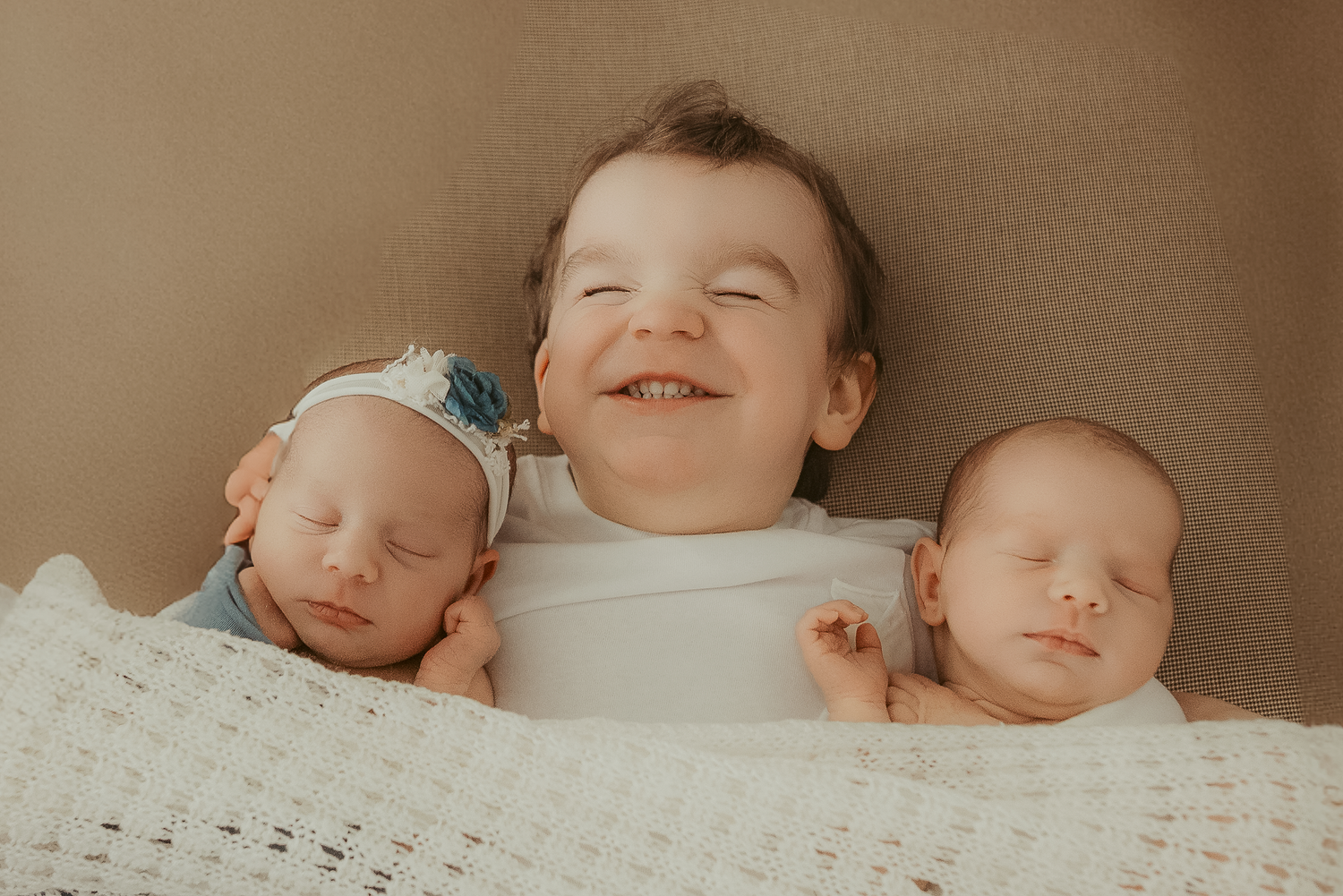 Smiling toddler posing with two sleeping newborn twins wrapped in white blankets for a studio photo.