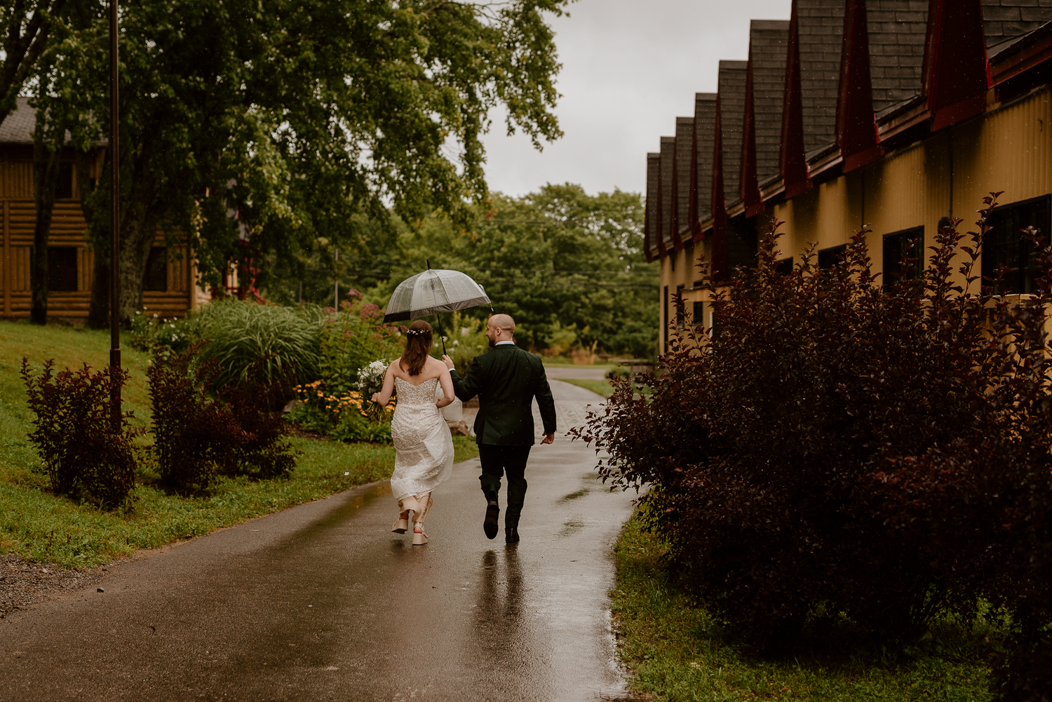 Couple walking in rain holding white umbrella along path by brick buildings.