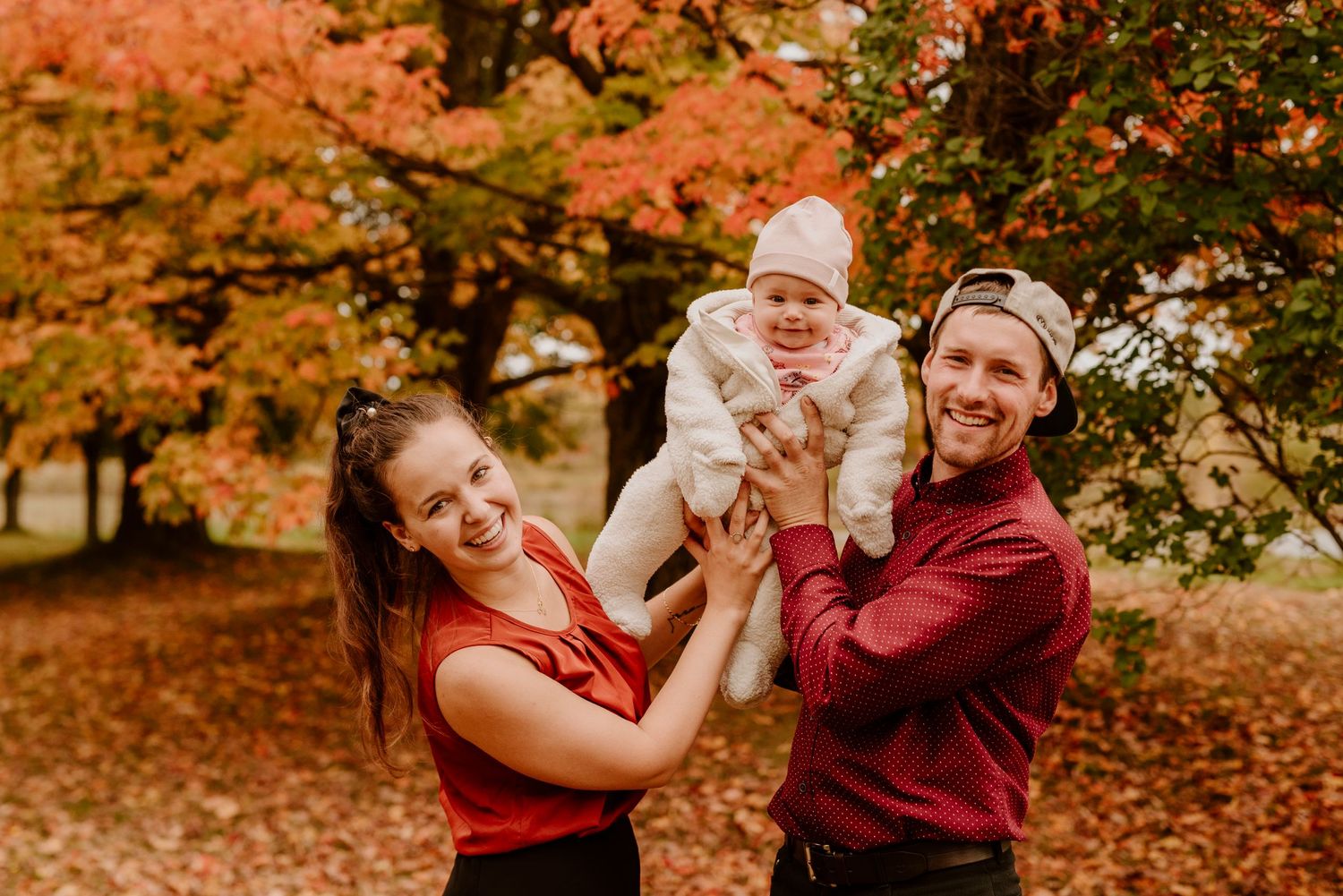 Une photo spontanée d'un couple et leur bébé, tous souriants, dans un décor automnal chaleureux.