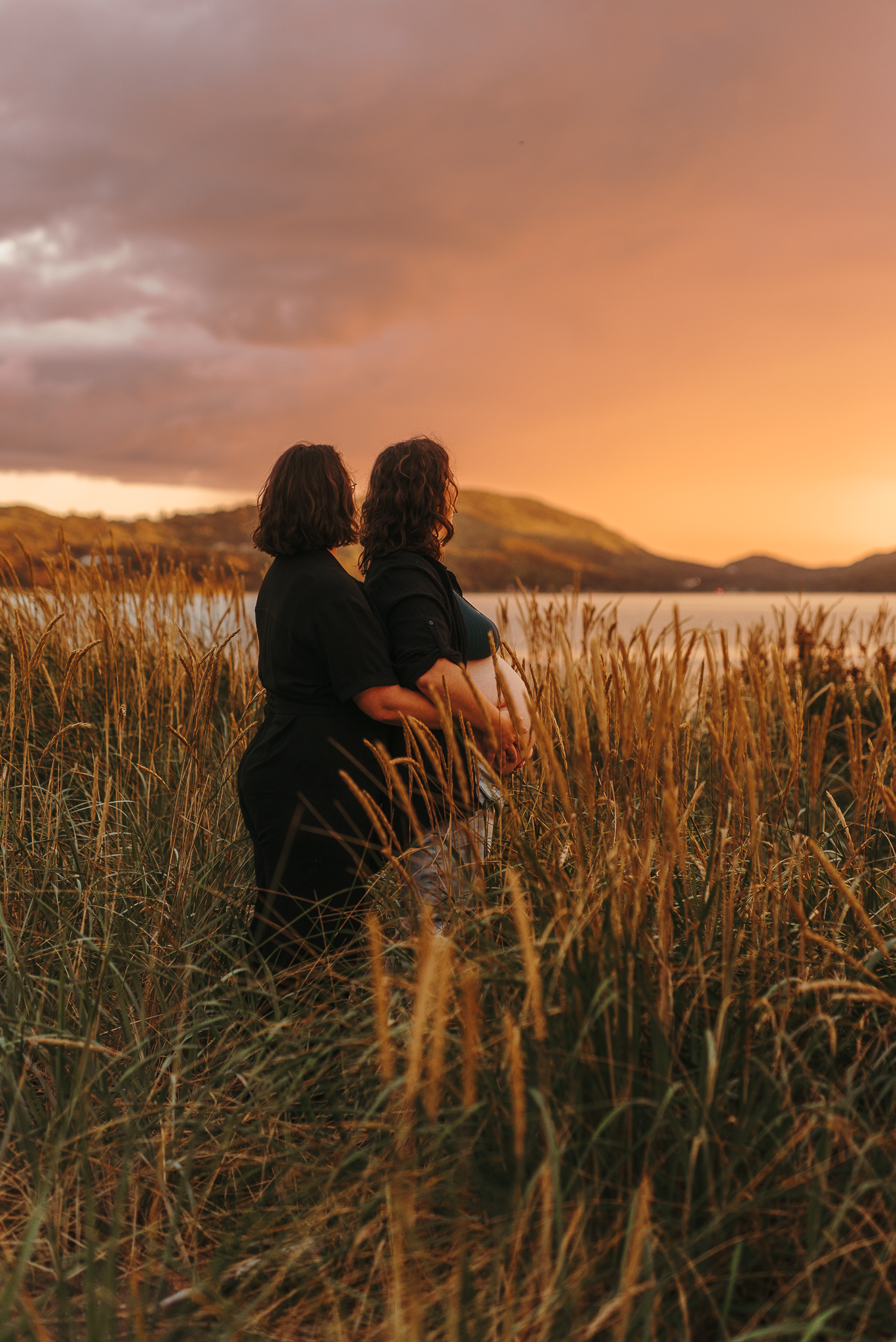 Two people embrace in tall golden grass at sunset, with a dramatic orange sky and serene lake in the background.