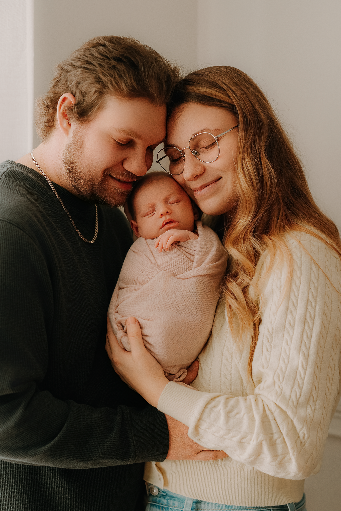 New parents tenderly cradle their sleeping newborn baby, heads touching, sharing a loving family moment.