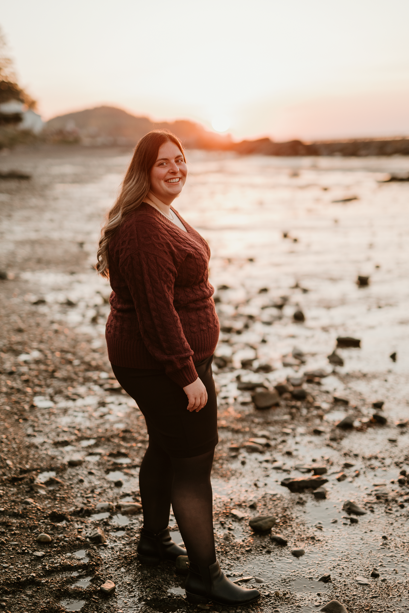 Pregnant woman in burgundy sweater smiling at sunset on rocky beach, golden hour glow behind her.
