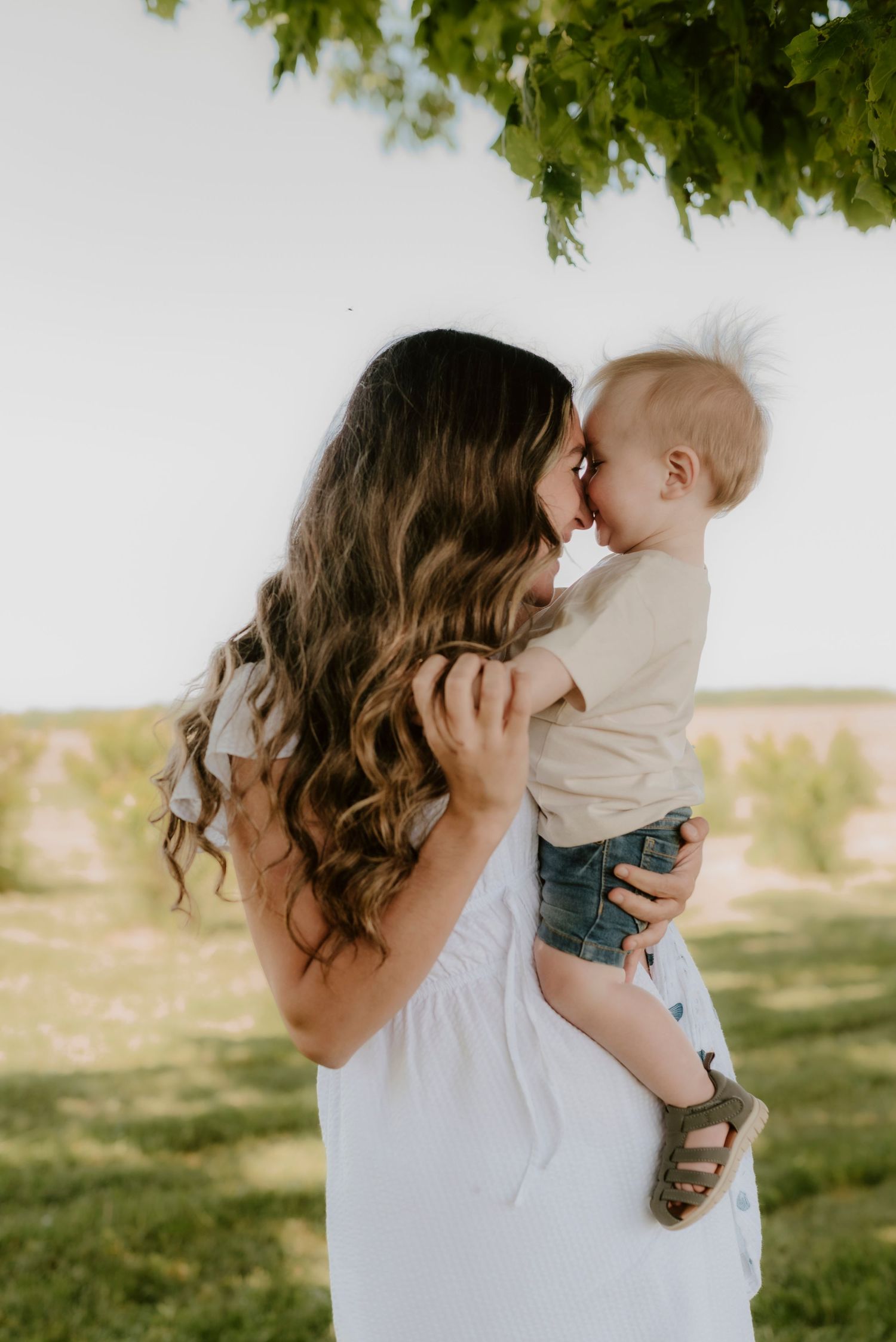 A tender moment between parent and baby in a sun-dappled outdoor setting with green foliage overhead.