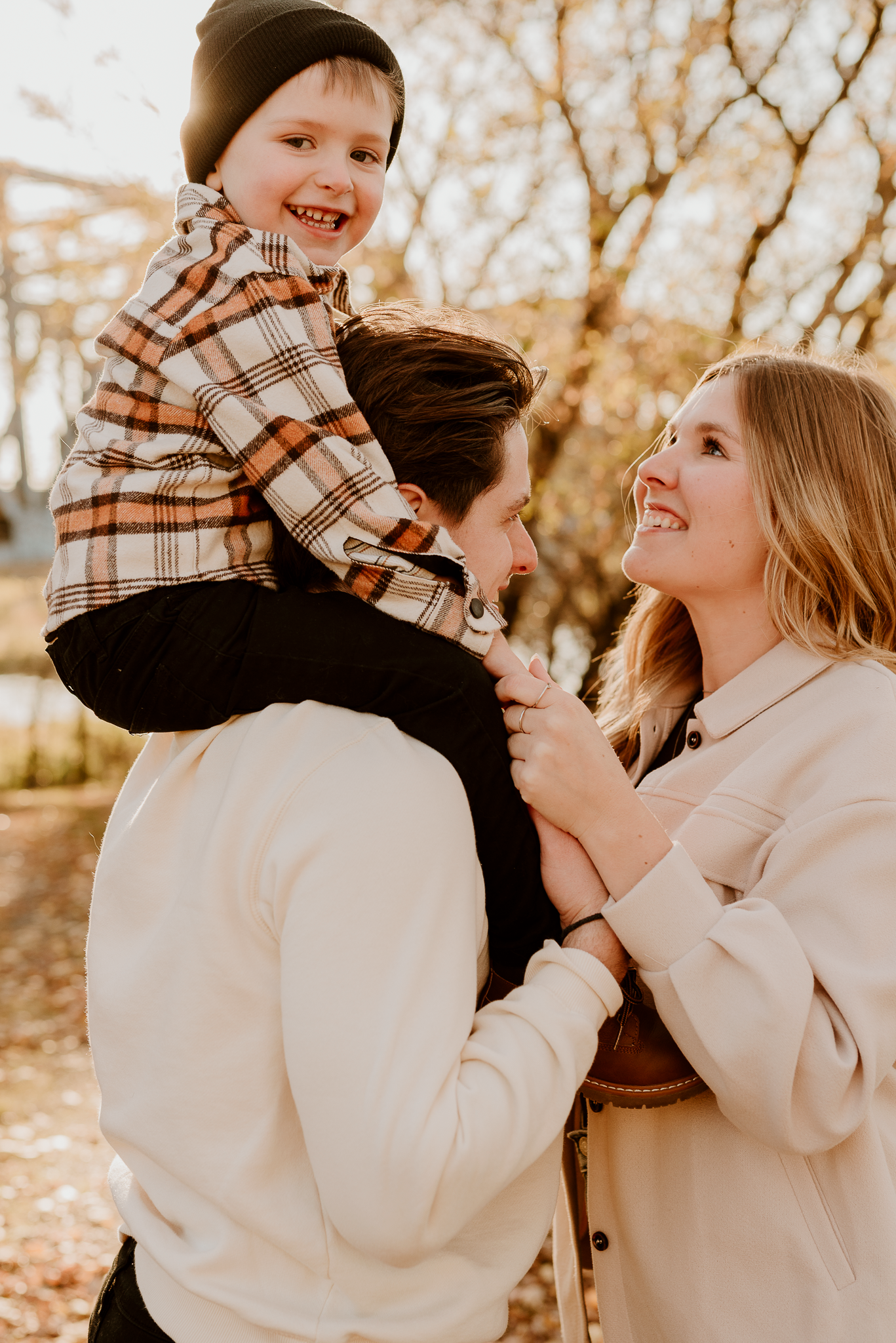 A joyful family of three enjoys a sunny autumn day outdoors, with a laughing toddler perched on dad's shoulders.