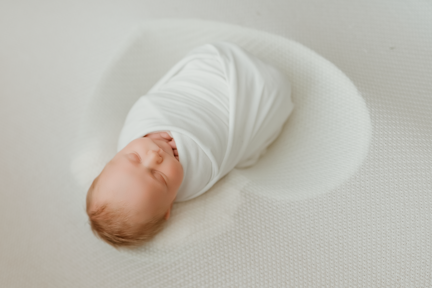 Newborn baby swaddled in white wrap sleeping peacefully on a soft white blanket.