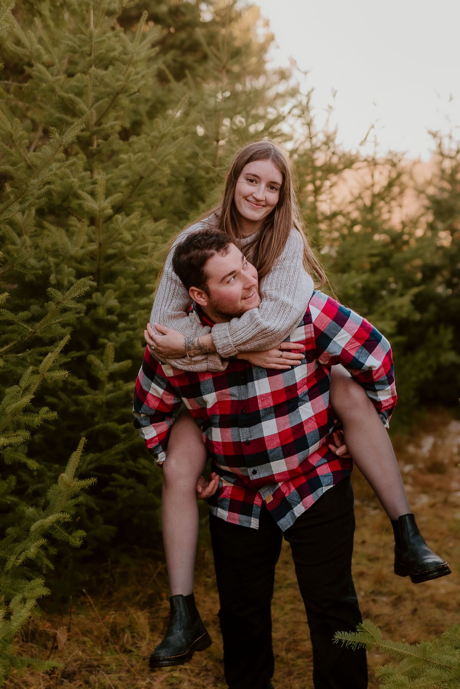 A couple poses at a Christmas tree farm, woman giving piggyback ride smiling among lush green pine trees.