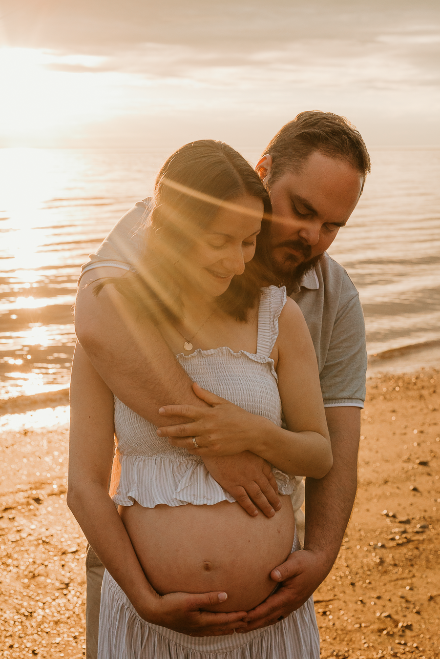 Couple tenderly embracing at sunset beach, man holding pregnant woman's belly by golden ocean waters.