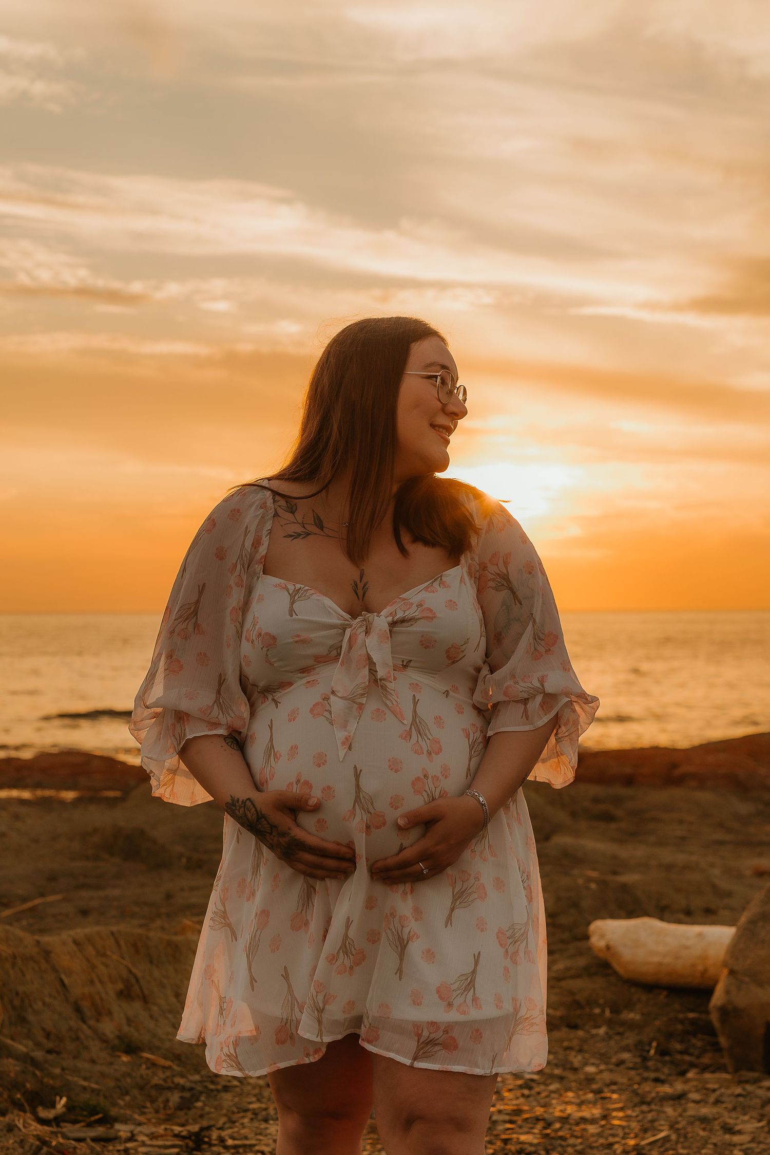 Pregnant woman in floral dress cradling baby bump at golden sunset by the ocean.